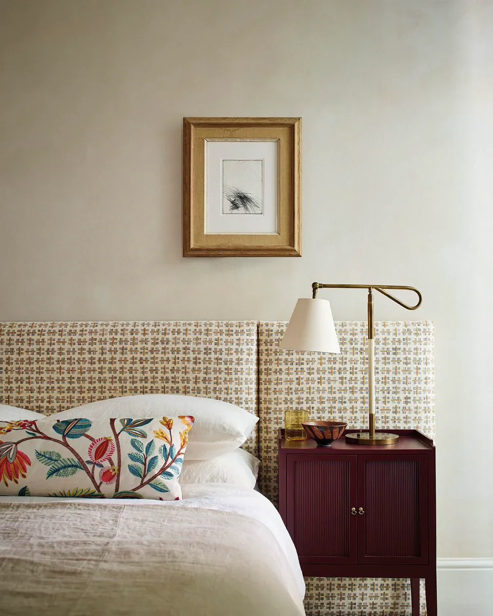 A bedroom corner with a bed, a colorful embroidered pillow, a beige headboard, a burgundy bedside table, a gold and white adjustable lamp, a glass of yellow liquid, a bowl, and wall art above the headboard.