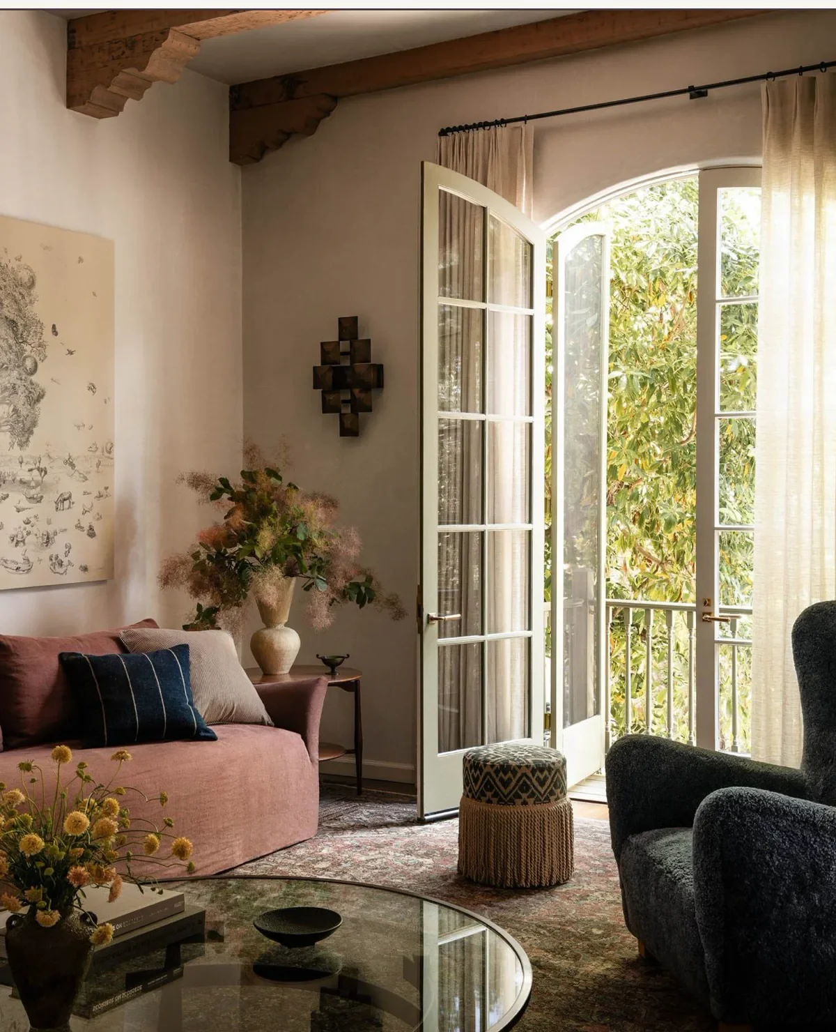 A cozy living room with open French doors leading to a balcony and green foliage outside. The room features a pink sofa with pillows, a floral arrangement in a white vase, a round glass coffee table, and a patterned ottoman. Light-colored curtains hang beside the doors, and wooden beams are visible on the ceiling.