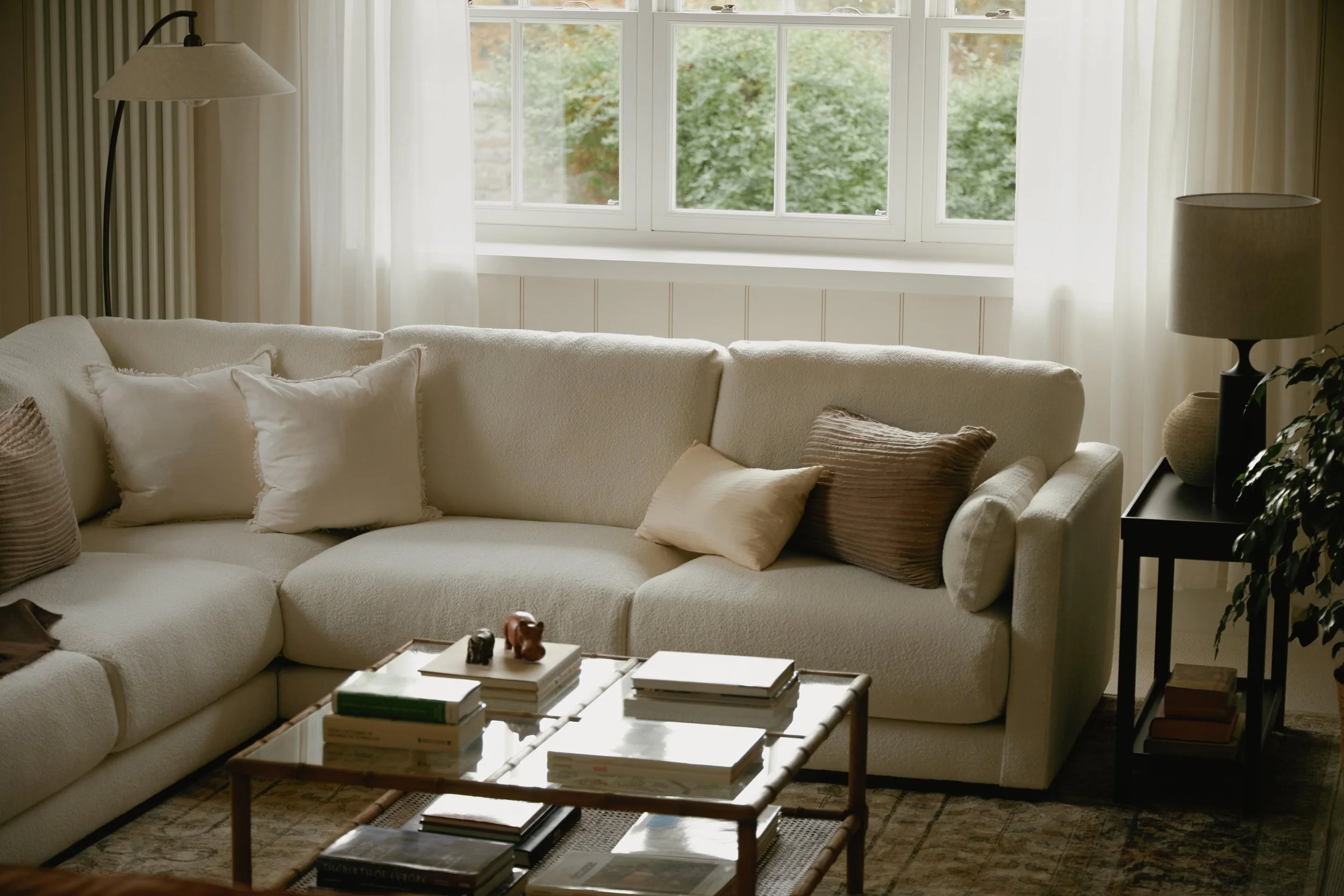 Living room with a large cream-colored sectional sofa, decorative pillows, a coffee table with books and small figurines, a window with white curtains, a floor lamp, a side table with a lamp, and some plants.