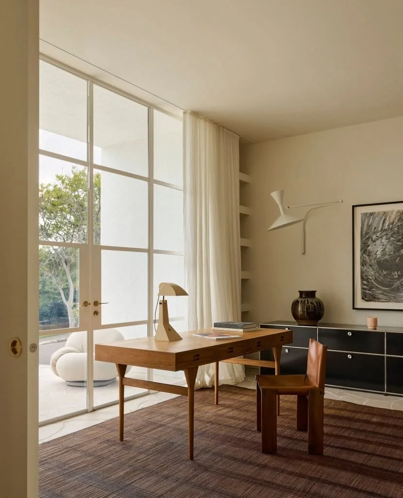 A modern, minimalist home office with a wooden desk, leather chair, white desk lamp, black sideboard, and decorative items including a large vase and framed artwork. Floor-to-ceiling windows with white curtains overlook a patio and greenery outside.