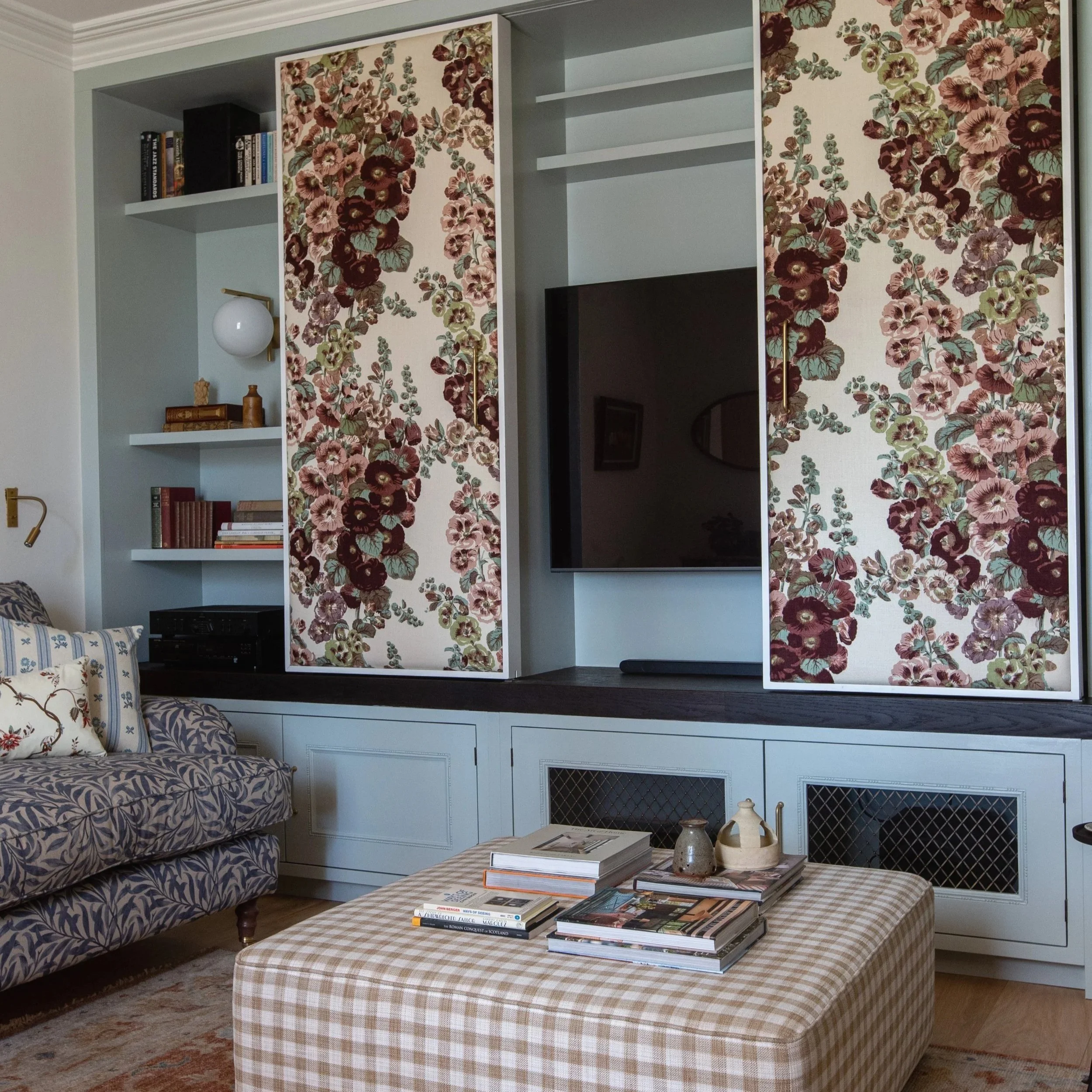 Living room with a white built-in shelf unit featuring floral wallpaper doors, a navy and white patterned sofa, a plaid ottoman with books and magazines, and a coffee table with decorative objects.