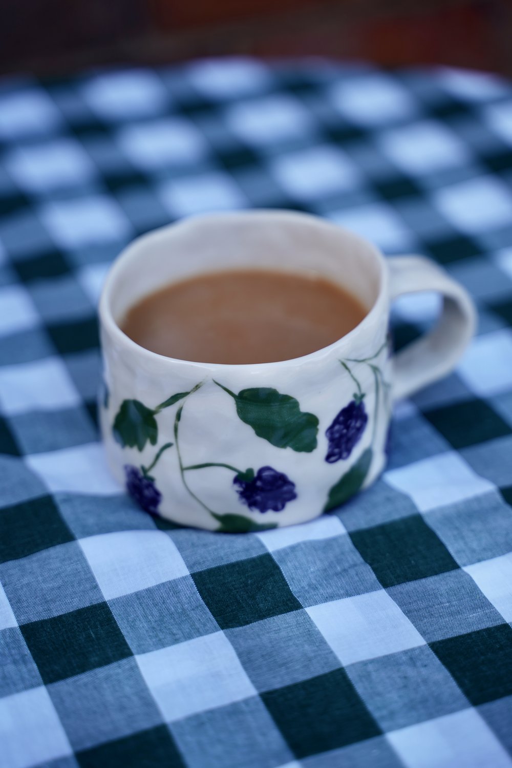 A handcrafted clay mug with painted blackberry pattern from Dill Homeware, shown on a gingham table cloth