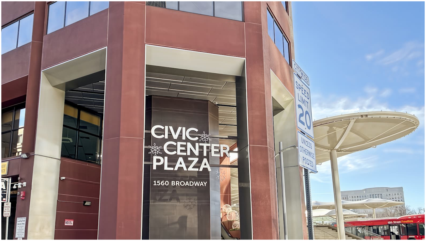 A photo of the red office building next to some patio umbrellas. The sign reads Civic Cener Plaza, 1560 Broadway.