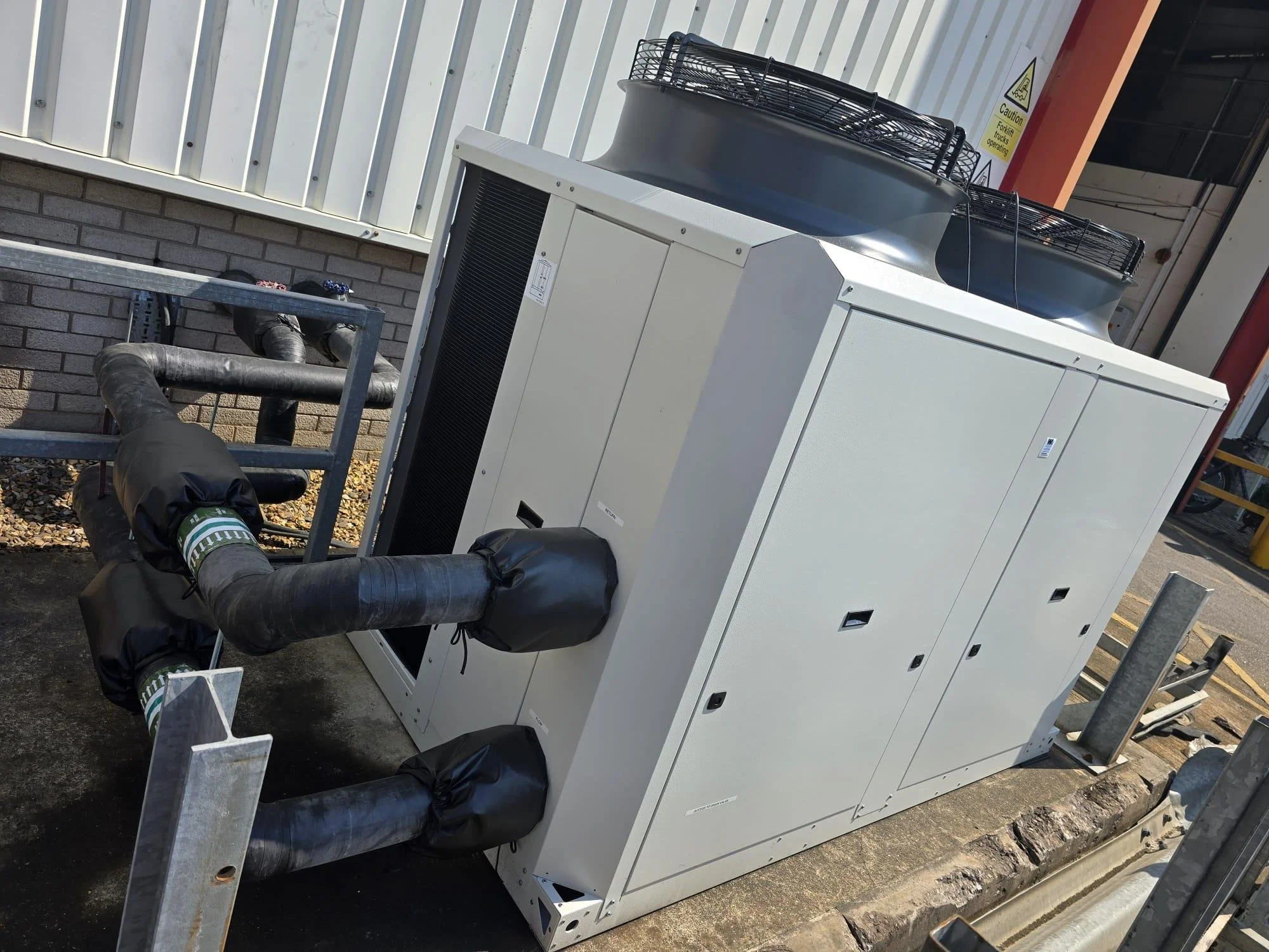Large white industrial HVAC unit with black ducts connected to it, installed outside near a brick wall. There is a caution sign on the wall nearby.