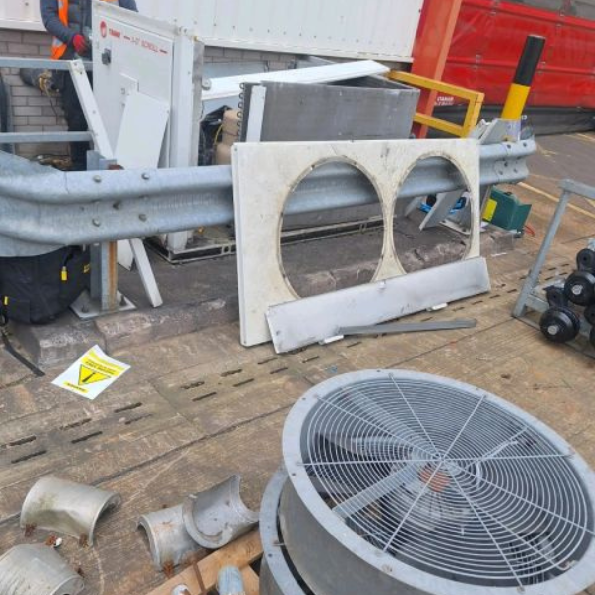 Disassembled parts of an air conditioning unit or industrial equipment on a wooden floor, including a large fan or vent cover, metal pipes, and various other metal and plastic components.