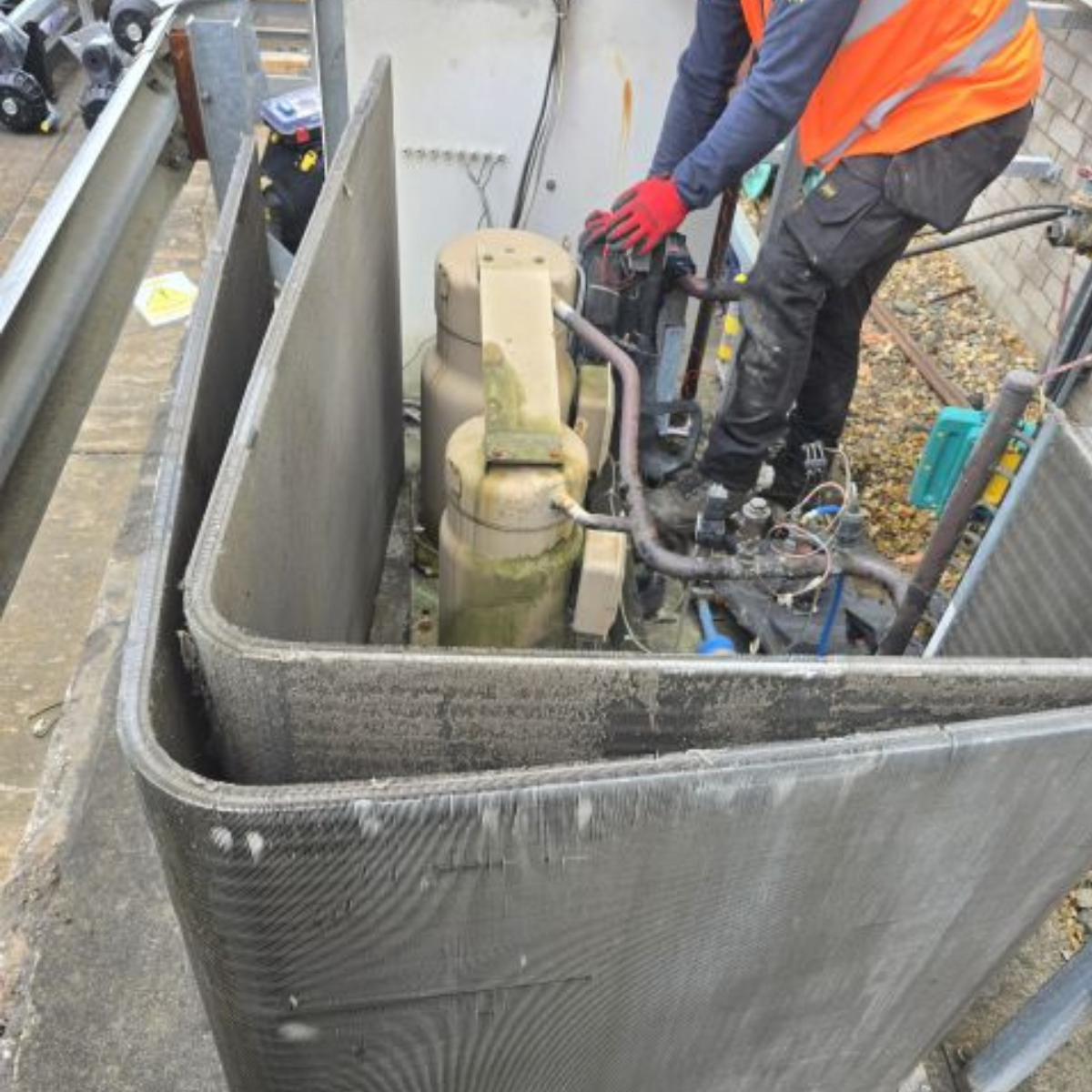 Construction worker wearing a safety vest and gloves working on HVAC equipment inside a construction site enclosed by a metal barrier.