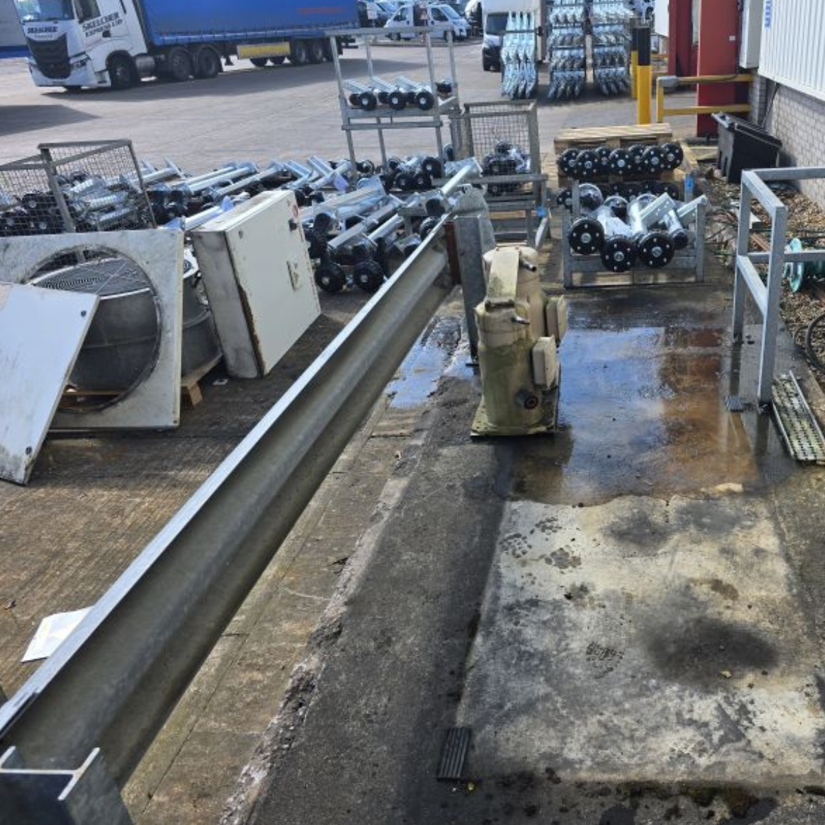 Outdoor industrial area with pipes, metal parts, and equipment scattered on wet concrete ground, adjacent to a parking lot with trucks and vehicles.