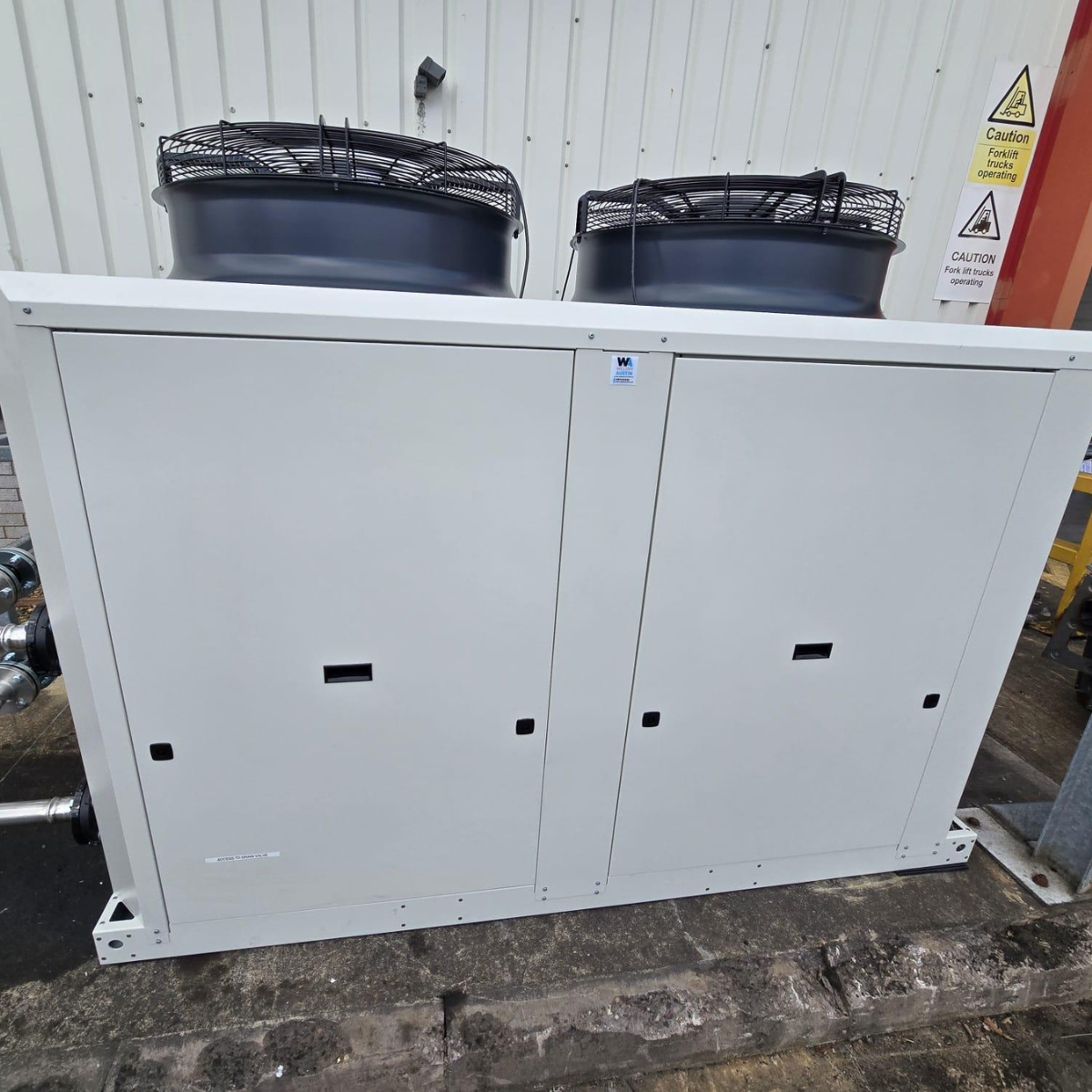 Large outdoor HVAC unit with two fan compartments on top, situated outside a building with white corrugated siding. Caution signs about forklift operation are visible nearby.