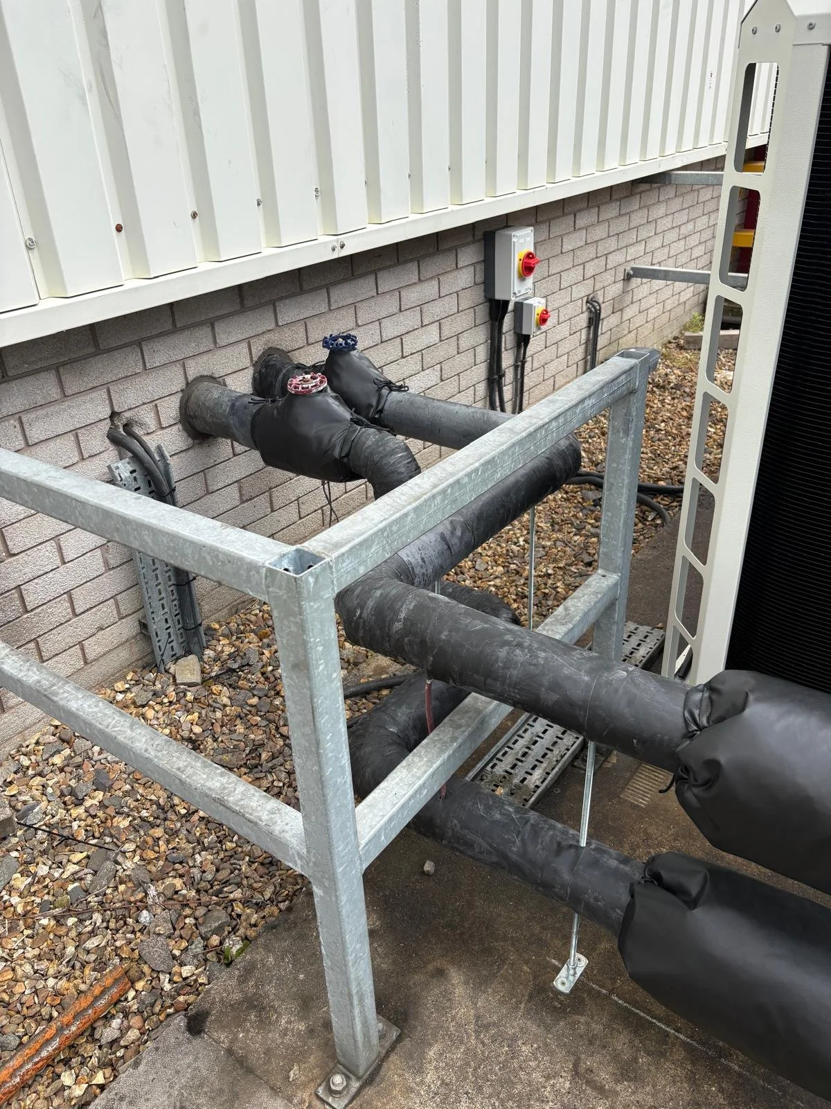 Outside view of an HVAC unit with insulated pipes wrapped in black foam, mounted on a metal frame against a brick wall. There are electrical disconnect switches on the wall behind the pipes.