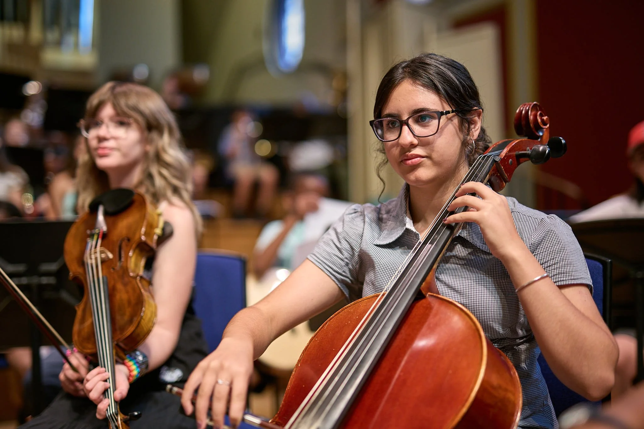 A young girl smiling whilst playing the cello, with a young violist in the background
