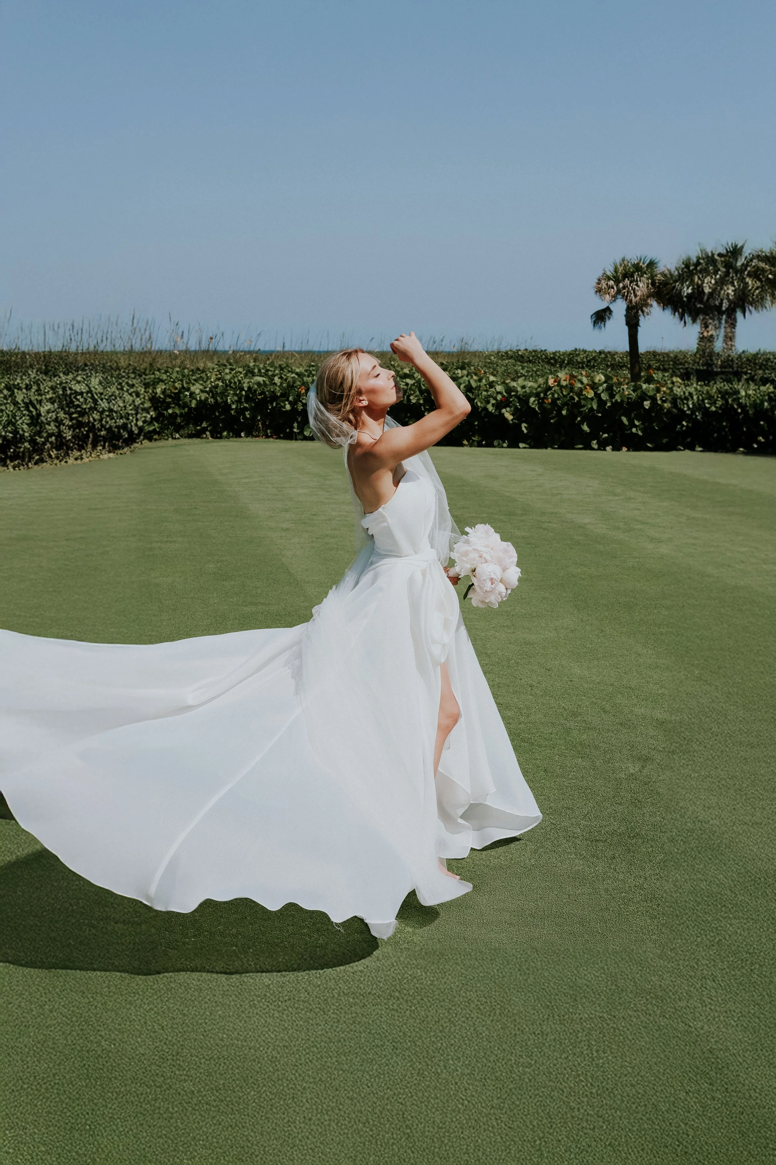 A bride wearing a white wedding dress, holding a bouquet, is standing on a green lawn outdoors under a clear blue sky. She is holding her veil and appears to be enjoying the moment.