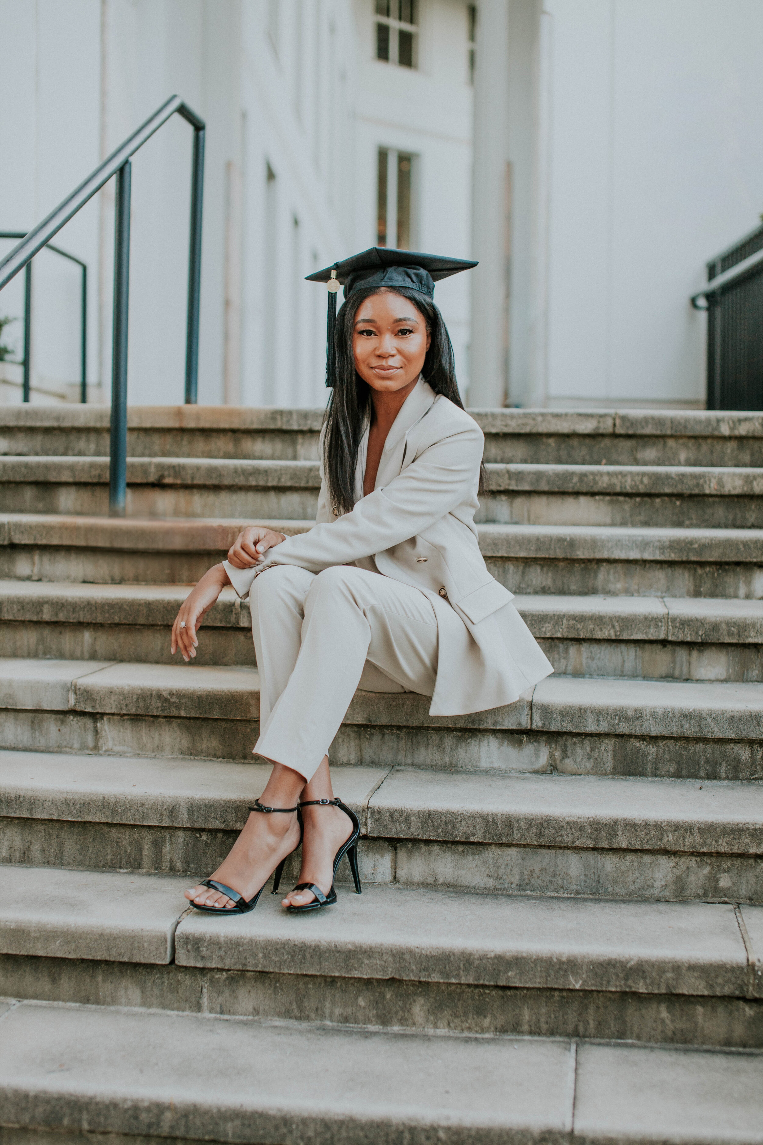 A person wearing a light-colored suit and graduation cap sitting on concrete steps, with black high heels, posing for a portrait in a city setting, Atlanta.