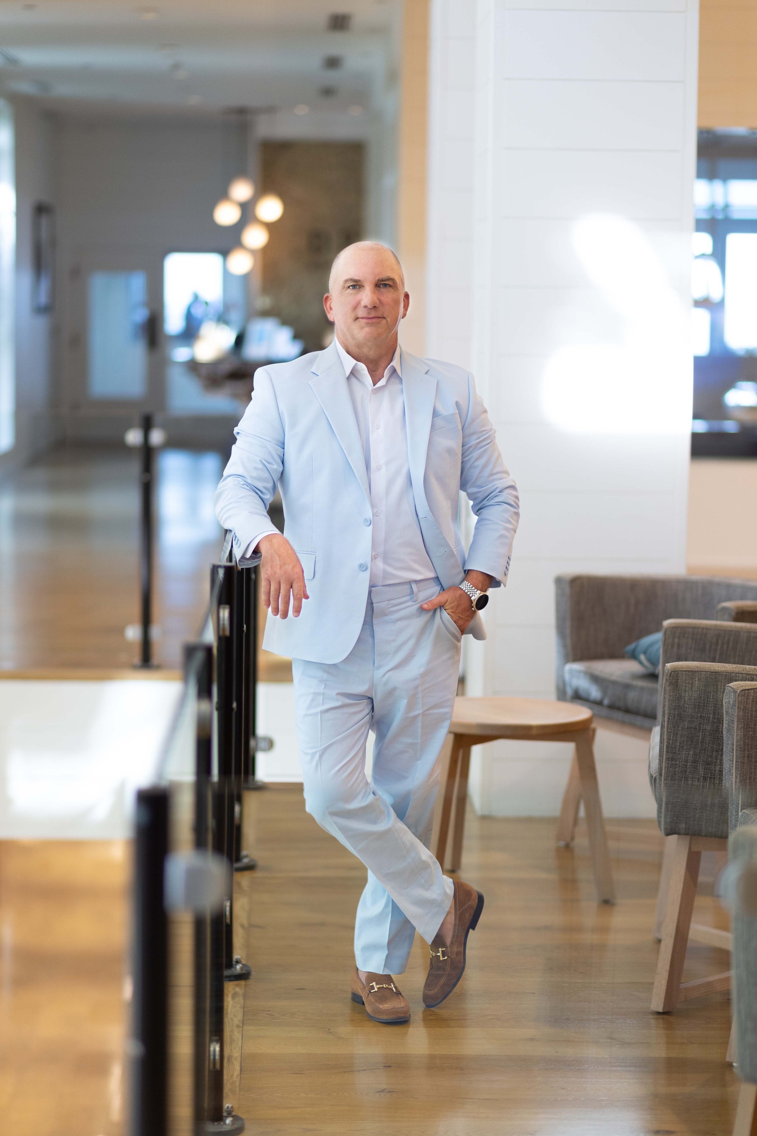 Man in light blue suit standing in a modern interior, leaning against a railing, with blurred background of seating area, lifestyle headshots.