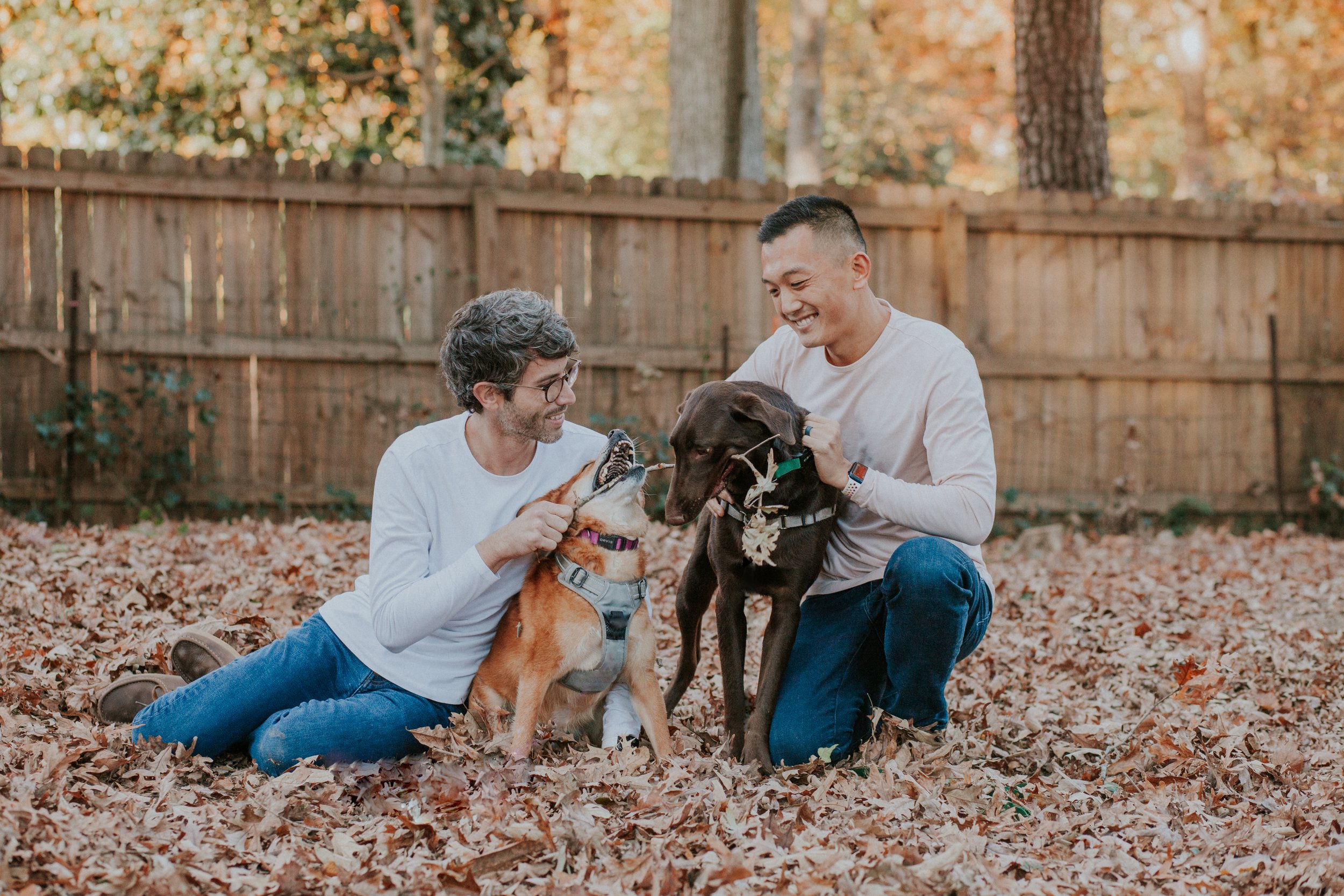 Two people sitting and kneeling in fallen leaves, petting two dogs in a backyard during autumn, captured in a family shoot in Atlanta.