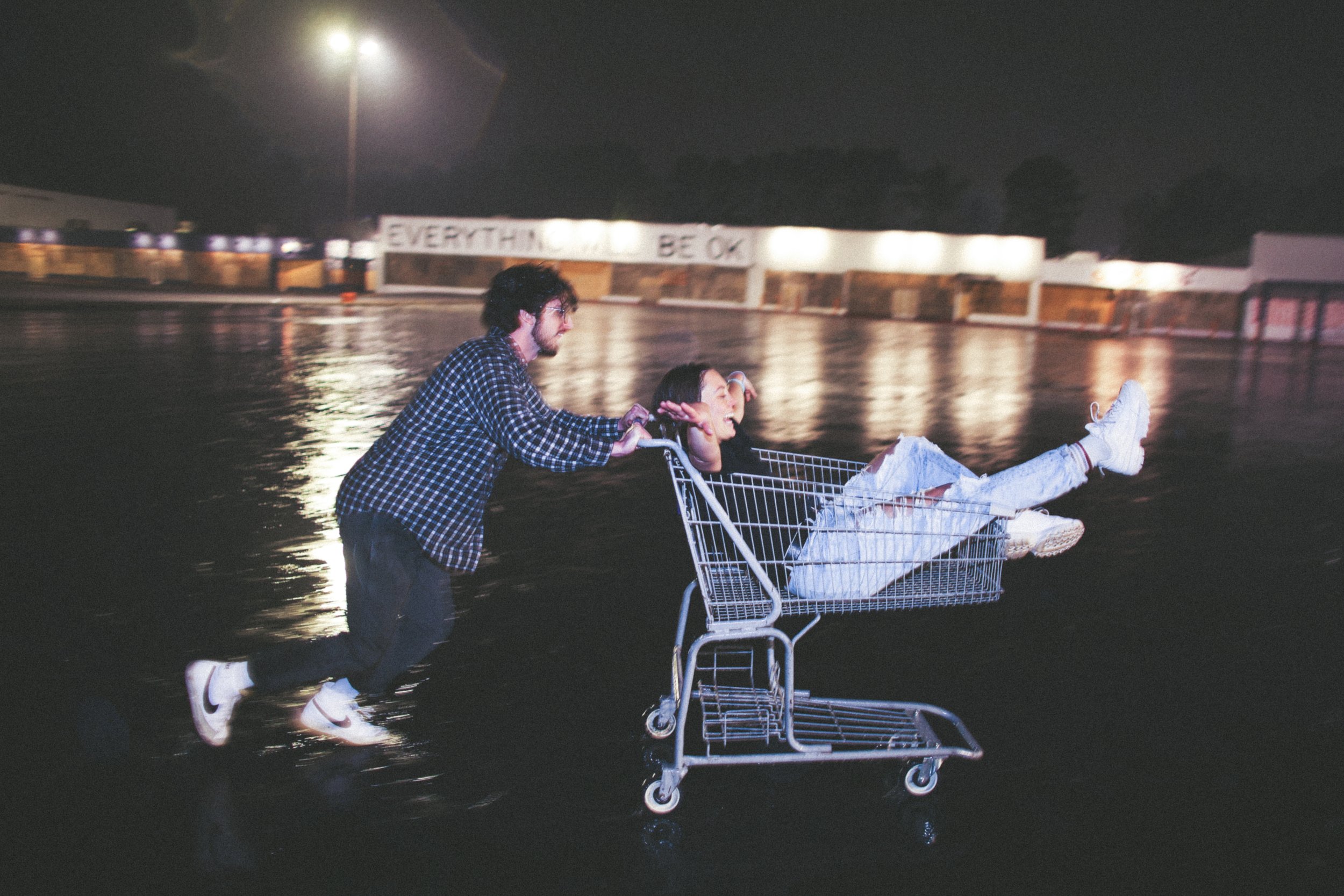 Two people having fun at night with a shopping cart in an empty parking lot, one pushing and one sitting inside.