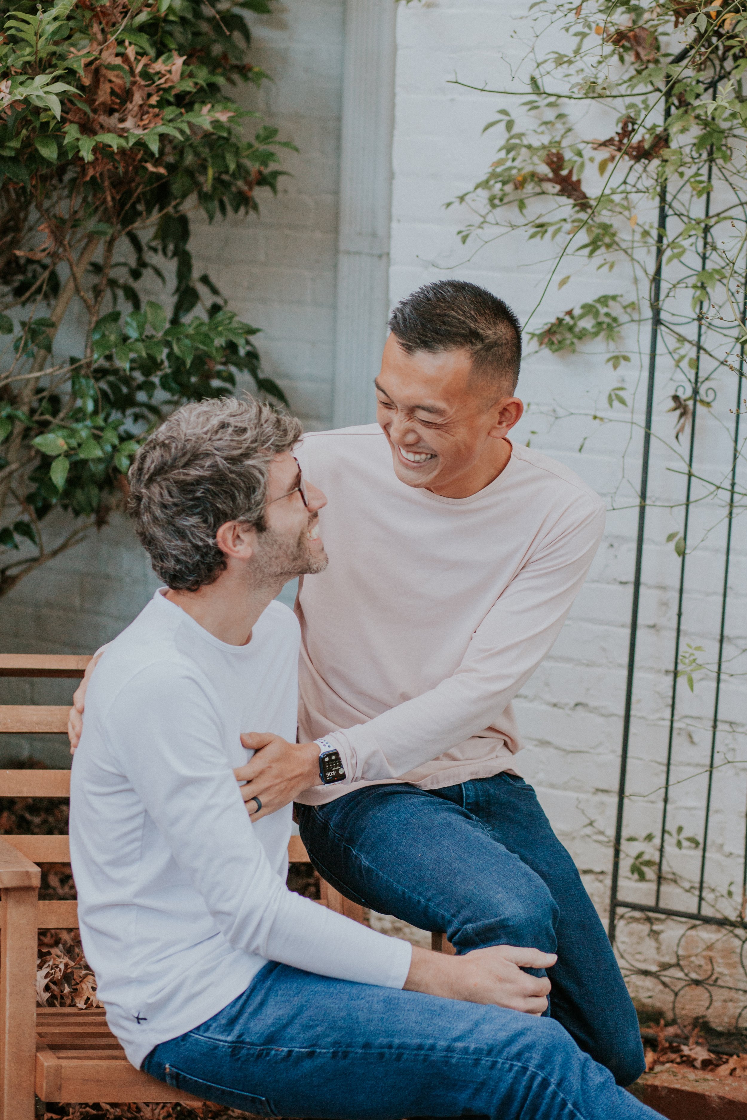 Two same-sex male couples sitting on a wooden bench outdoors, smiling and engaging with each other. The setting includes greenery and a white brick wall.