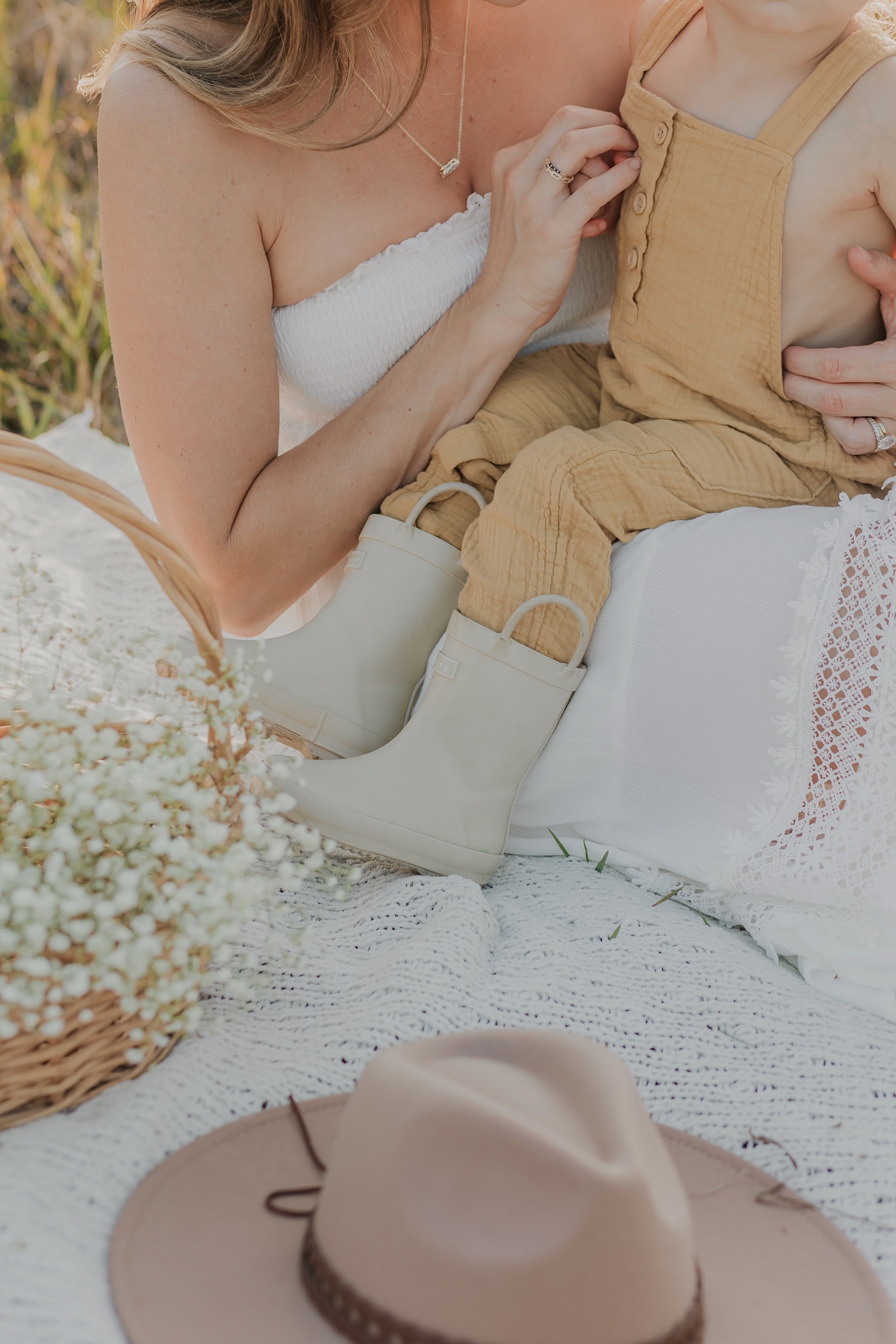 Woman and child sitting on a blanket outdoors, with a basket of flowers and a hat beside them. The child is wearing beige overalls and boots.