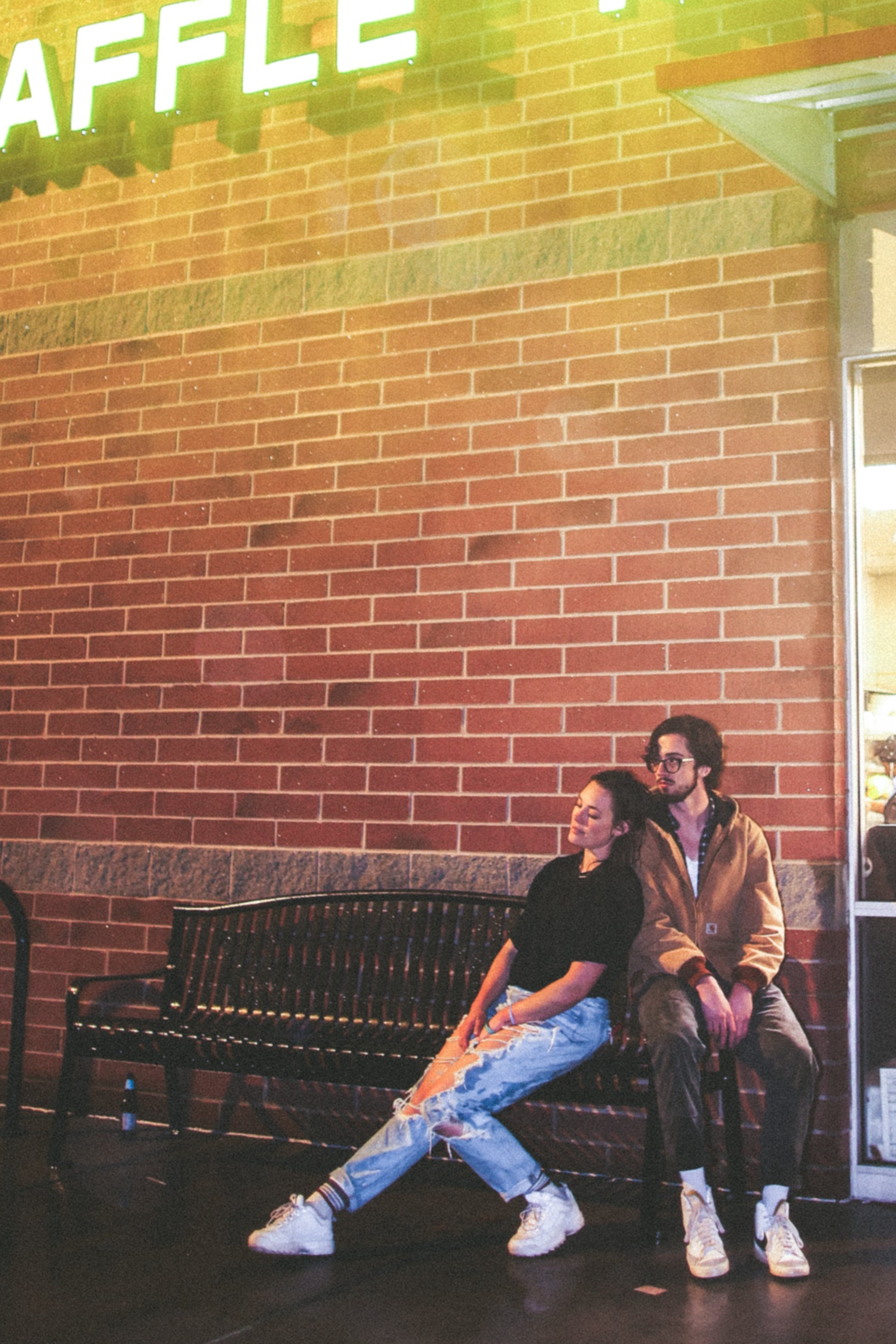 A couple sits on a bench outside a brick building under a neon light reading "WAFFLE." The woman with short hair is wearing ripped jeans and a black top, leaning affectionately on the man in a brown jacket and sunglasses.
