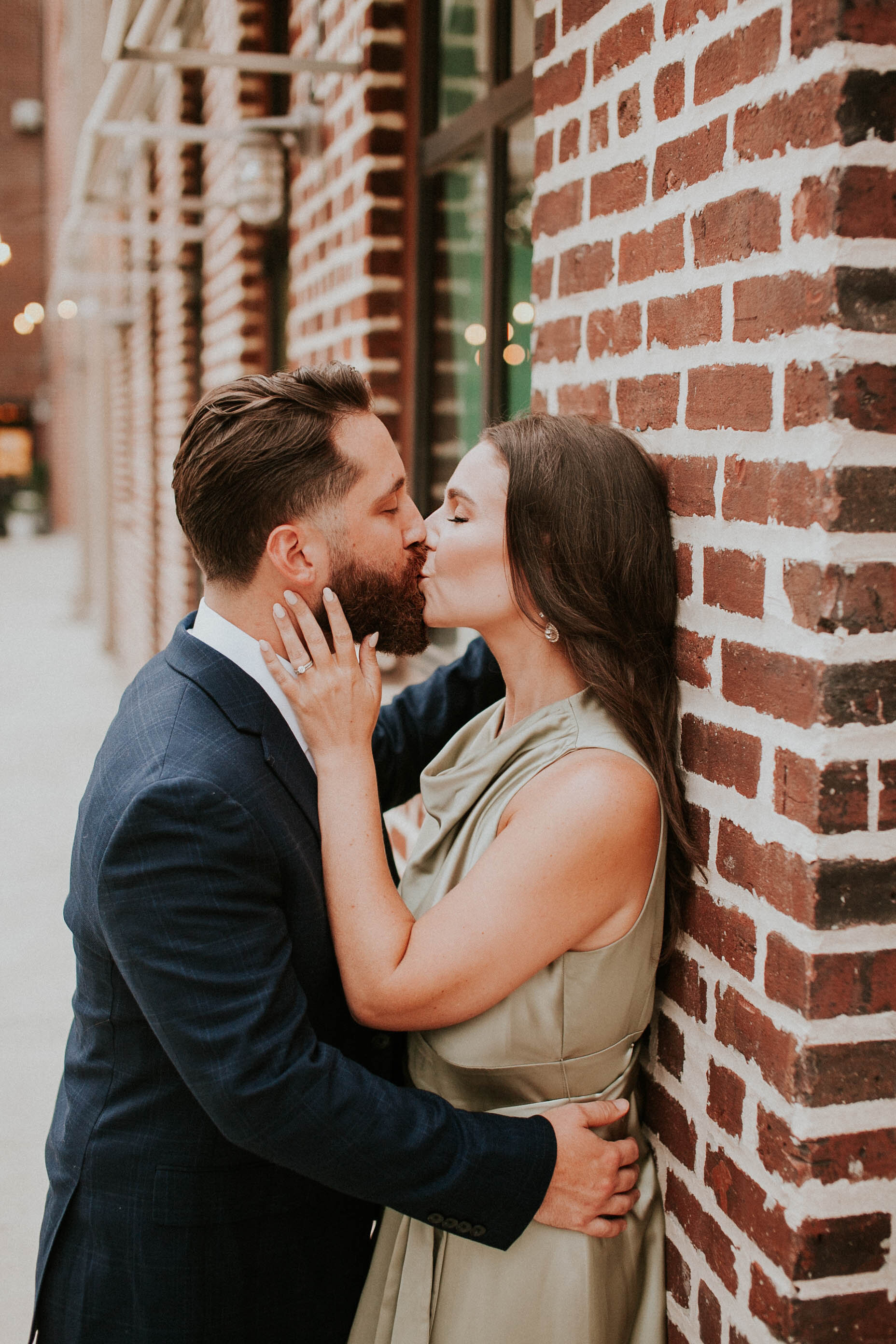 A couple kissing against a brick wall, with the woman wearing a green dress and the man in a suit.
