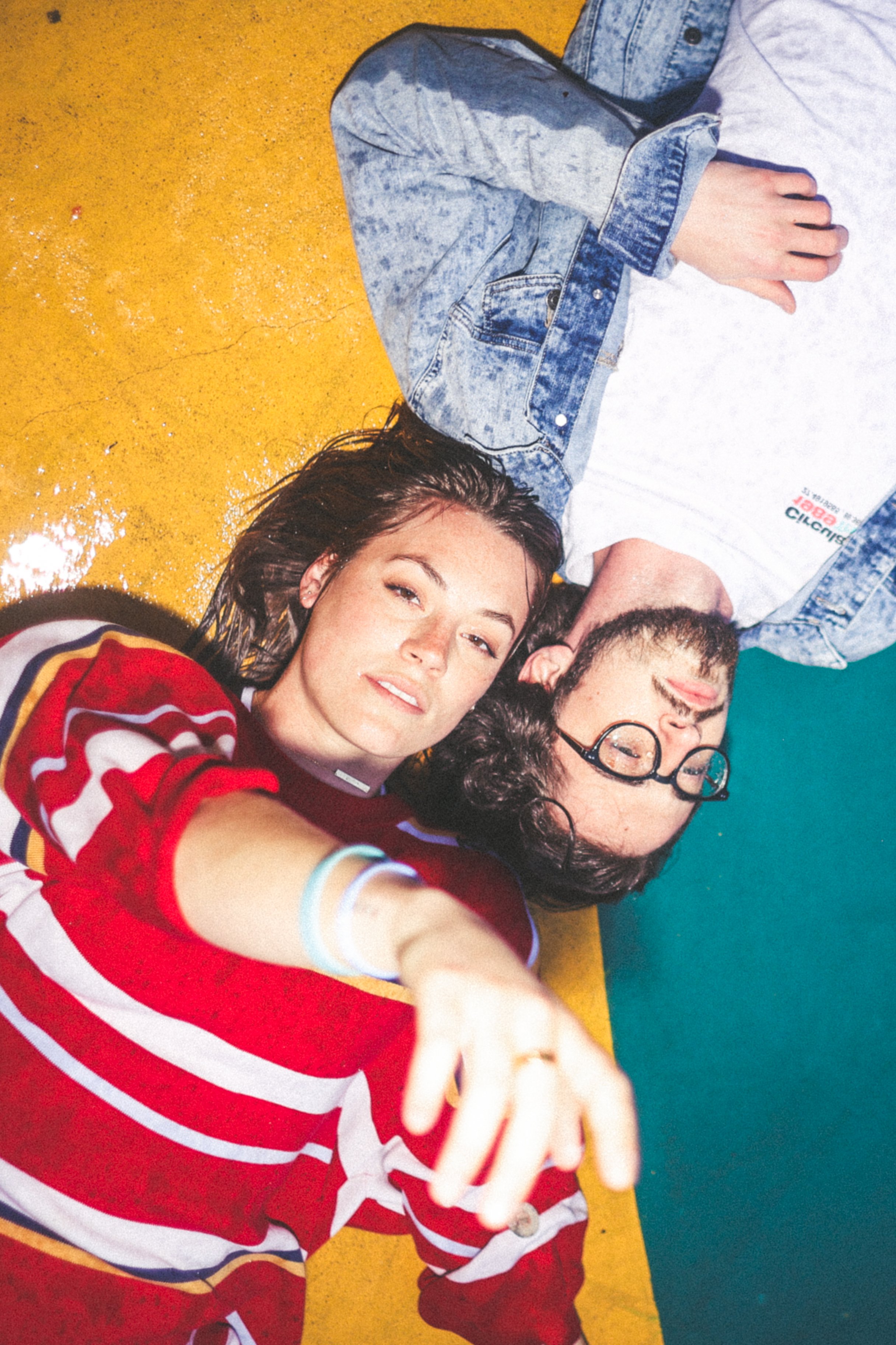 A woman in a red and white striped sweater lies beside a man in a denim jacket on a colorful floor. The woman is looking at the camera, while the man has his eyes closed.