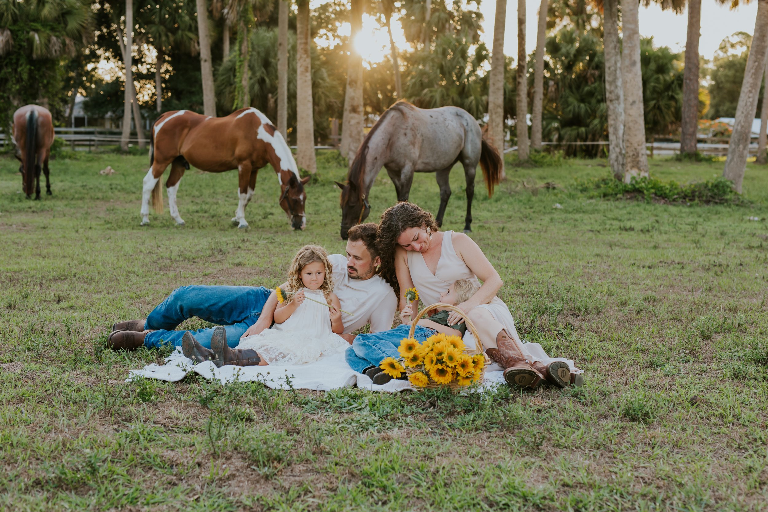 Family picnic in grassy Port Saint Lucie field with horses and sunflowers—man, woman in dress and cowboy boots, and two kids sitting on blanket—captured by South Florida family photographer