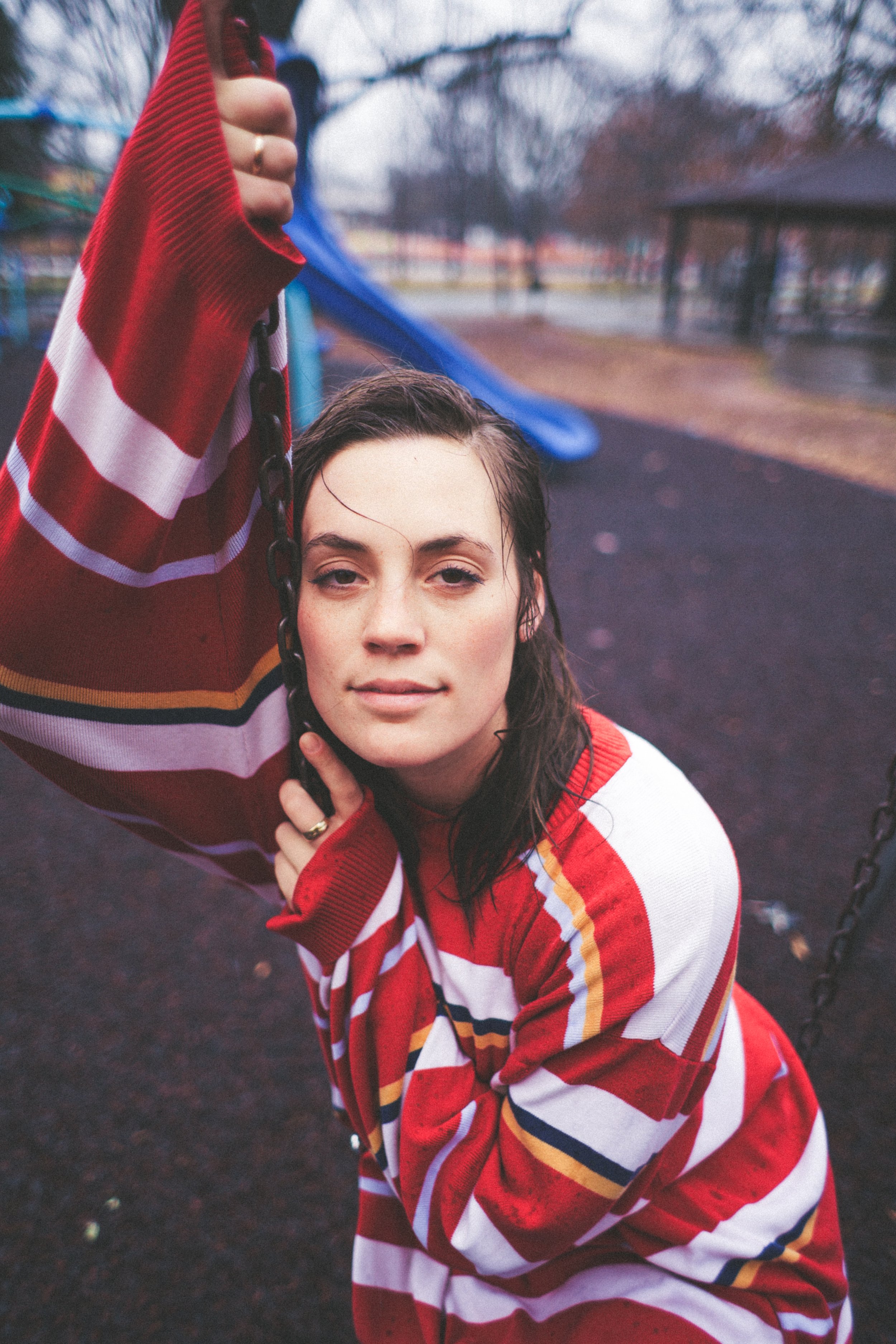Person wearing a striped red sweater holding a swing chain, standing in a playground on a rainy day.