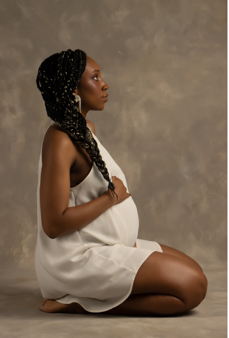 A pregnant woman wearing a white satin dress with long braided hair with blonde highlights, kneeling on the floor with a neutral expression, against a neutral background.
