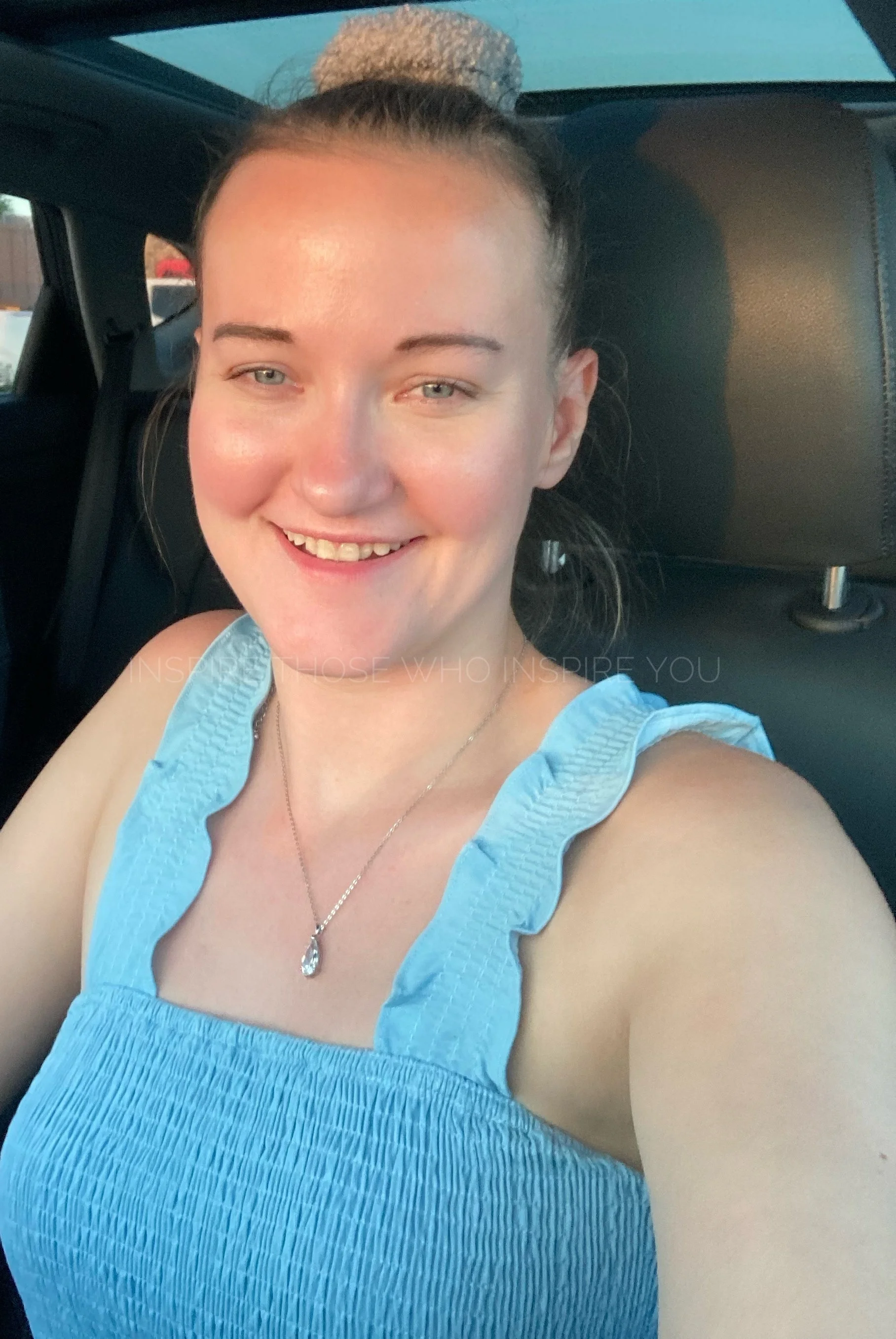 A smiling woman with light skin and blue eyes taking a selfie inside a car. She has her hair tied up in a bun with a hair scrunchie and is wearing a light blue sleeveless top with ruffled straps and a silver necklace with a teardrop-shaped pendant.
