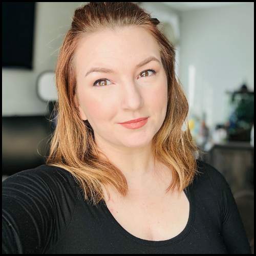 A young woman with light brown hair and fair skin taking a selfie indoors, wearing a black top with a room background.