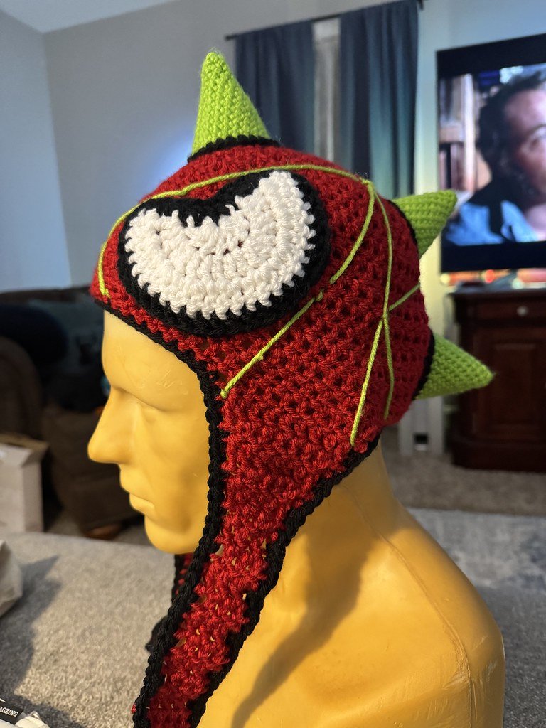 A handmade crochet hat resembling a cartoonish Spiderman, with green spikes, red body, black and white eye, and black outline, displayed on a yellow mannequin head.