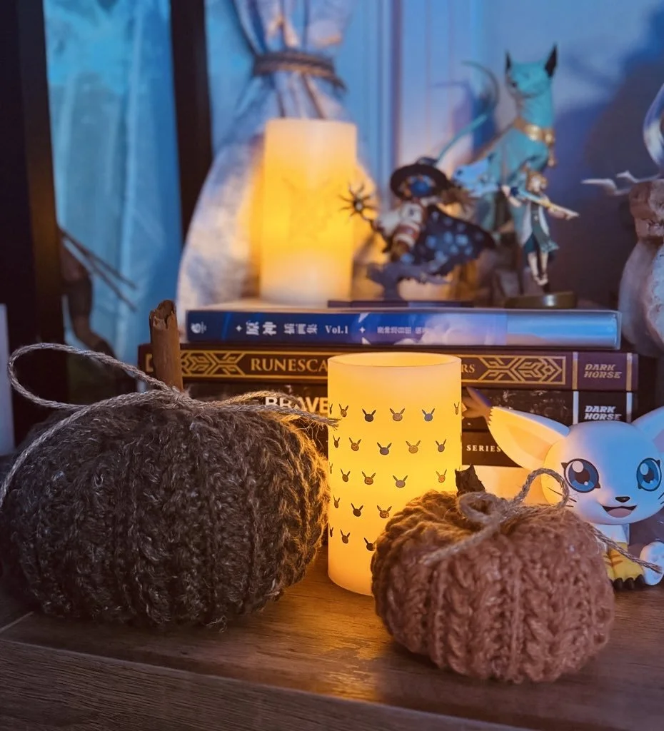 Decorative scene with two knitted pumpkins, a lit candle in a Bunny silhouette holder, books about dark horse games and runes, and figurines including a white creature with large eyes and a lengthy tail, set against a blue background with a lit candle and fantasy-themed decorations.