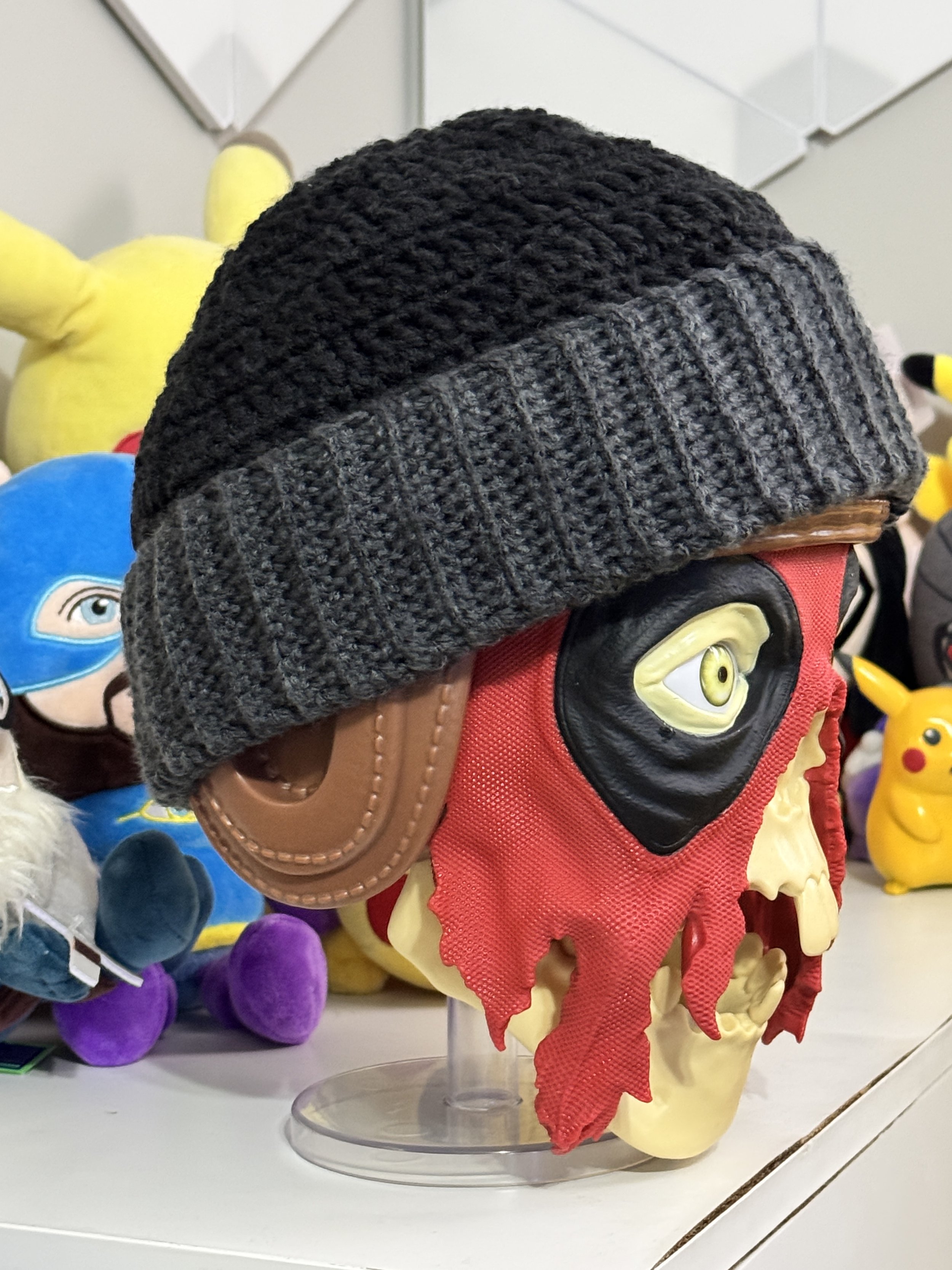 A display of a large skeleton mask with one eye, wearing a gray knit beanie, surrounded by plush toys of various characters.