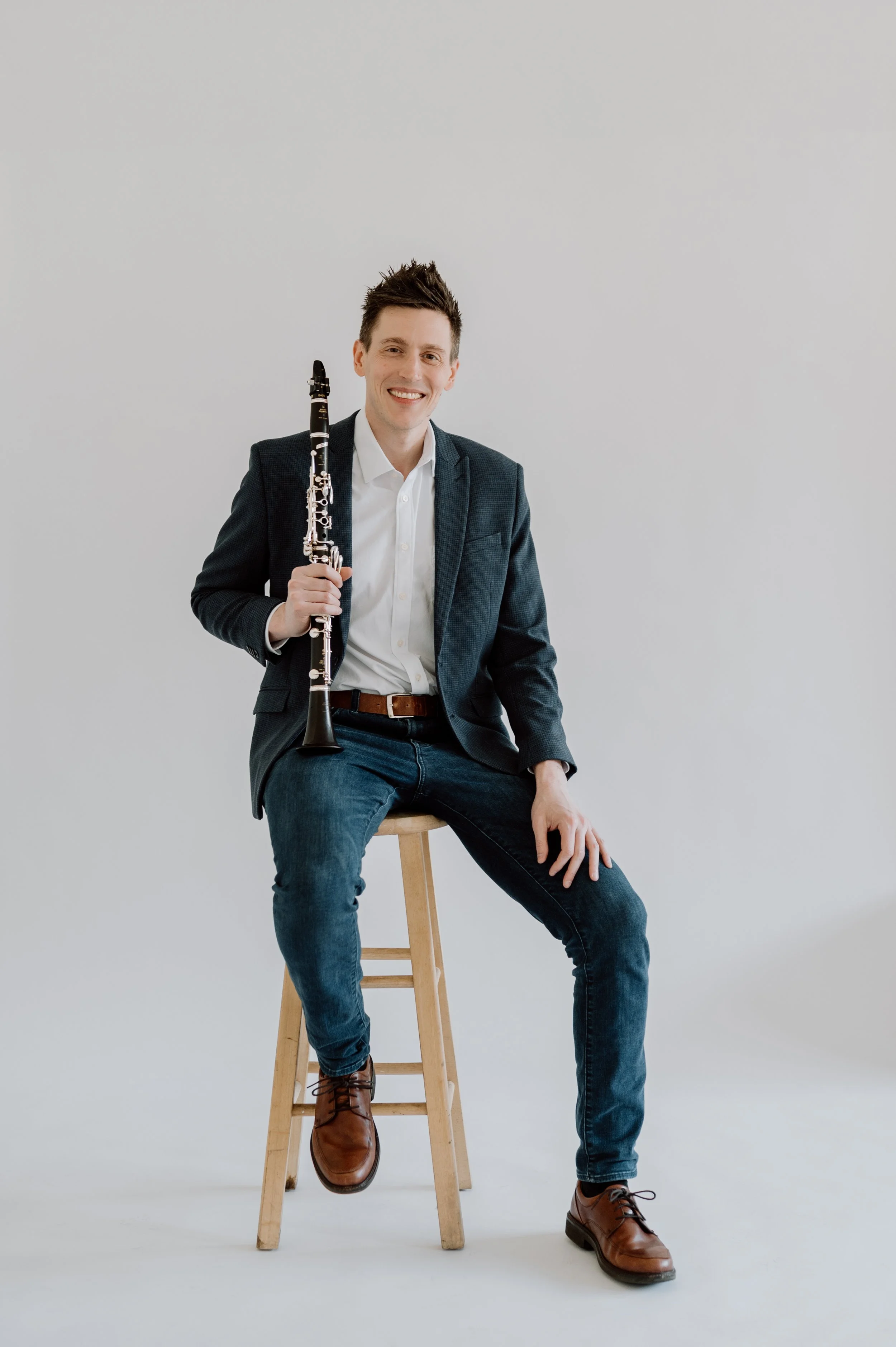 A man sitting on a wooden stool, holding a clarinet, smiling, wearing a white shirt, dark blazer, jeans, and brown leather boots, against a plain light gray background.