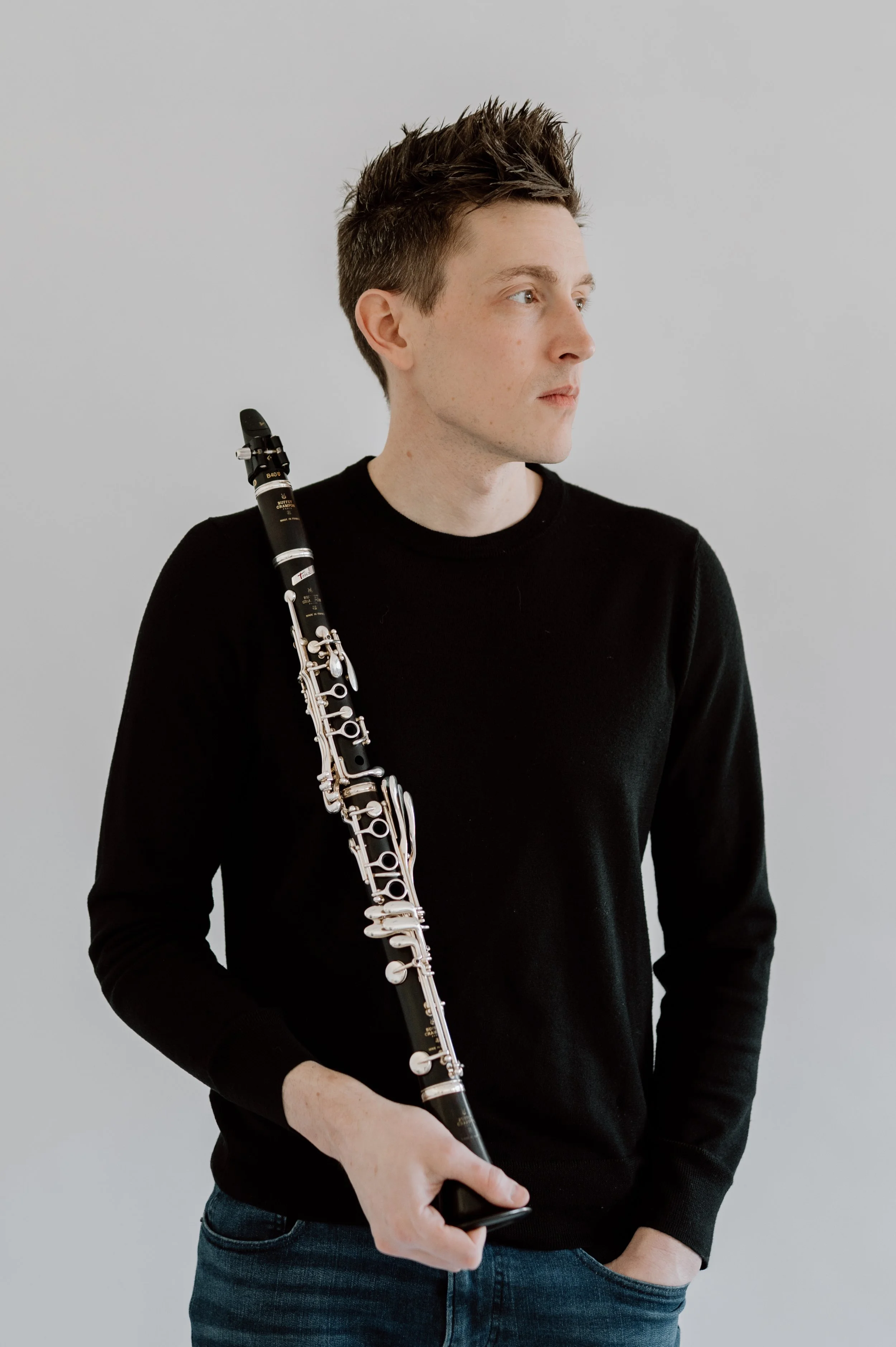 A young man with short spiky brown hair, wearing a black long sleeves shirt, holding a black soprano saxophone, and standing with one hand in his pocket against a plain light gray background.