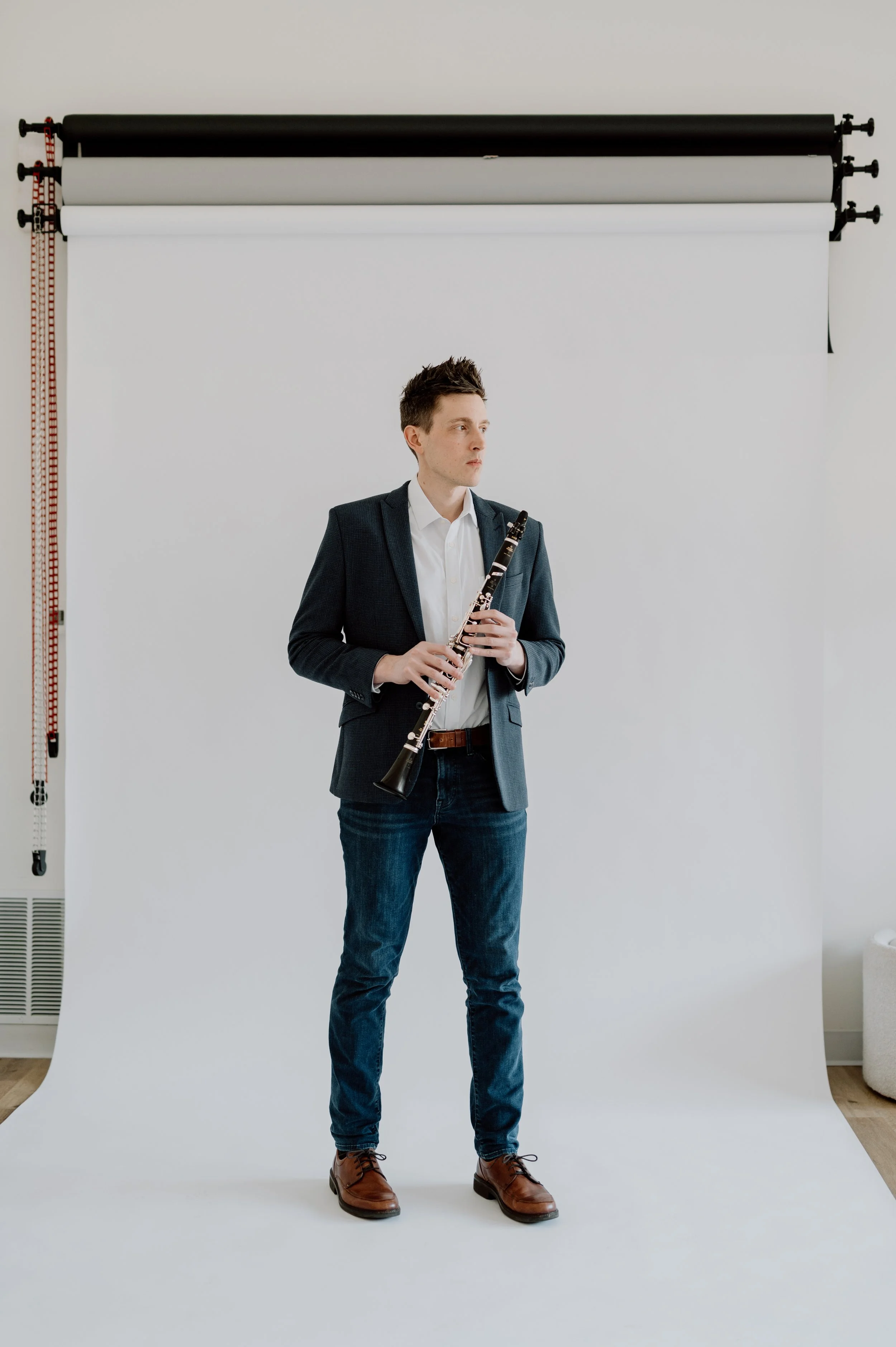 A young man in a grey blazer, white shirt, and jeans holding a clarinet, standing in front of a plain white backdrop in a photography studio.