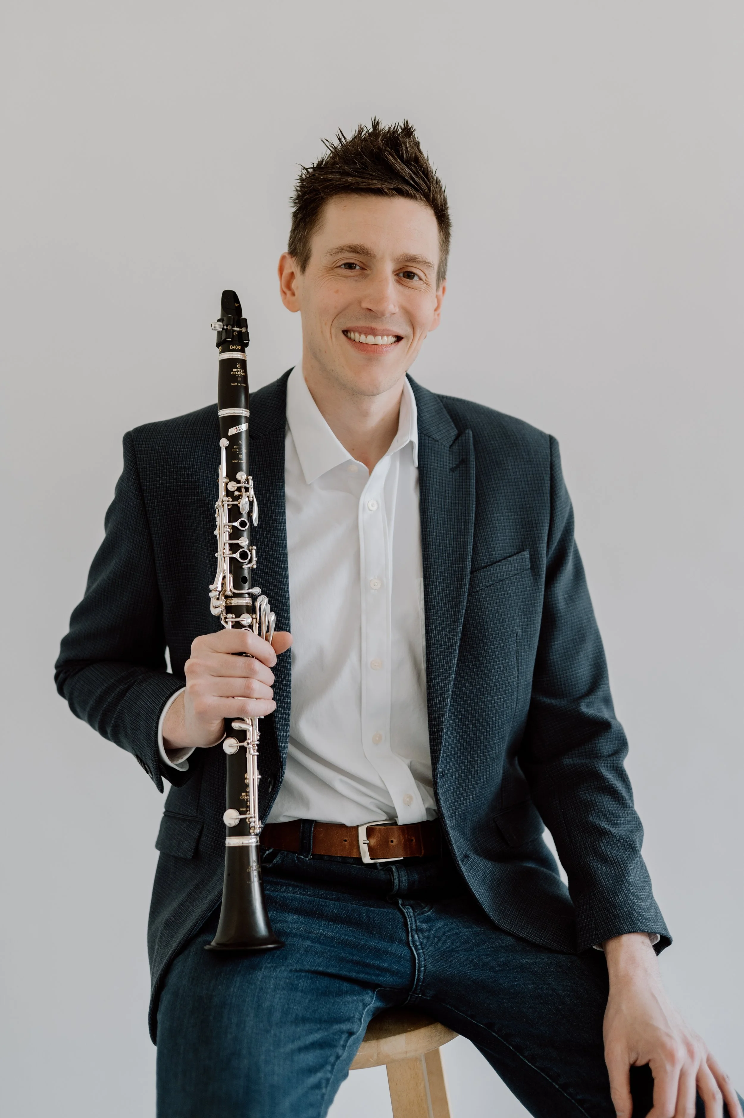 A smiling man with styled brown hair, wearing a dark checkered blazer, white shirt, and a brown belt, holding a black clarinet, seated on a wooden stool against a plain gray background.
