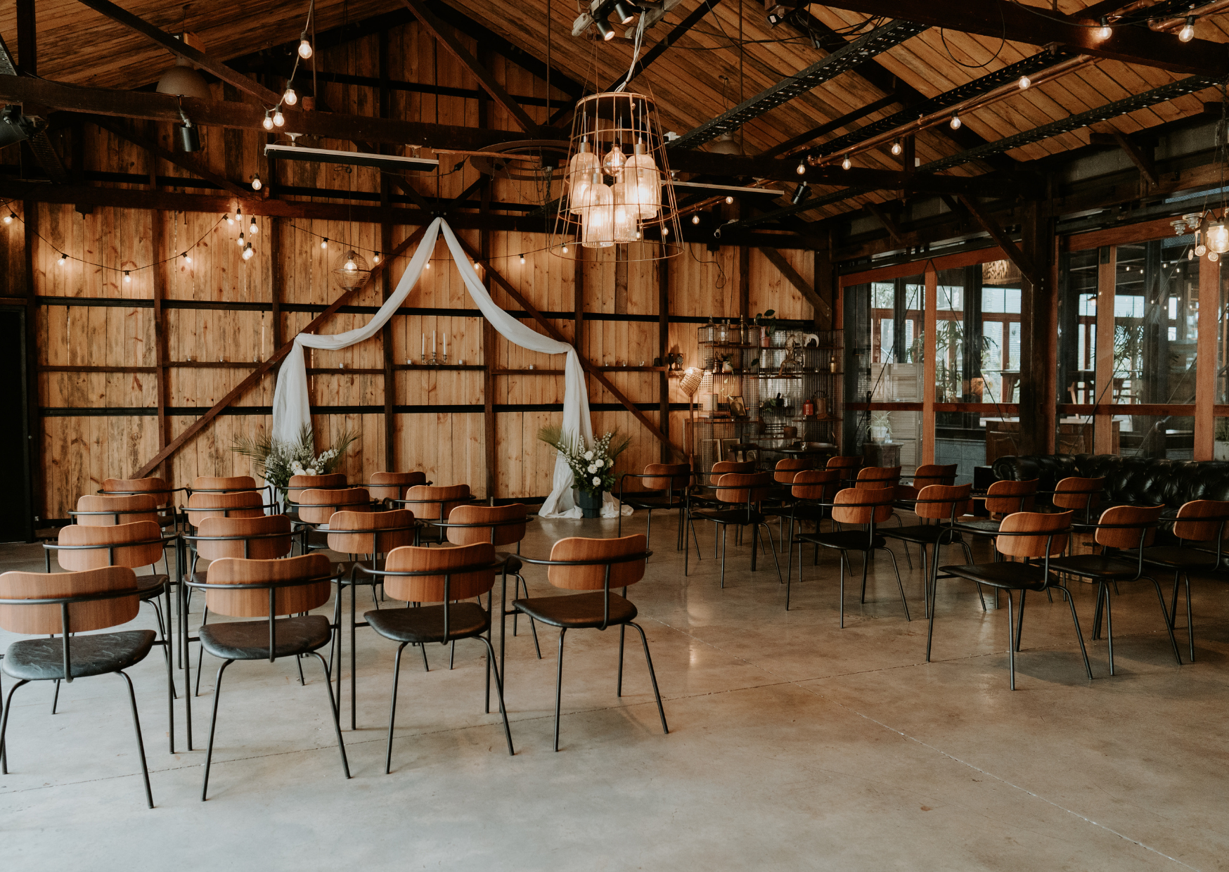 Empty rustic event space with string lights, wooden walls, chairs and stools arranged in semi-circle for a wedding ceremony, potted plants, and vintage furniture.