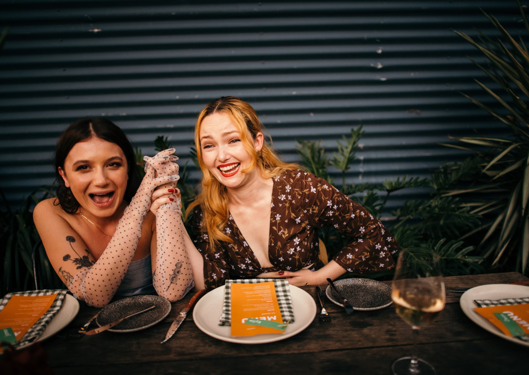 Two women sitting at a dinner table, smiling and laughing, with their hands clasped together, having fun together.