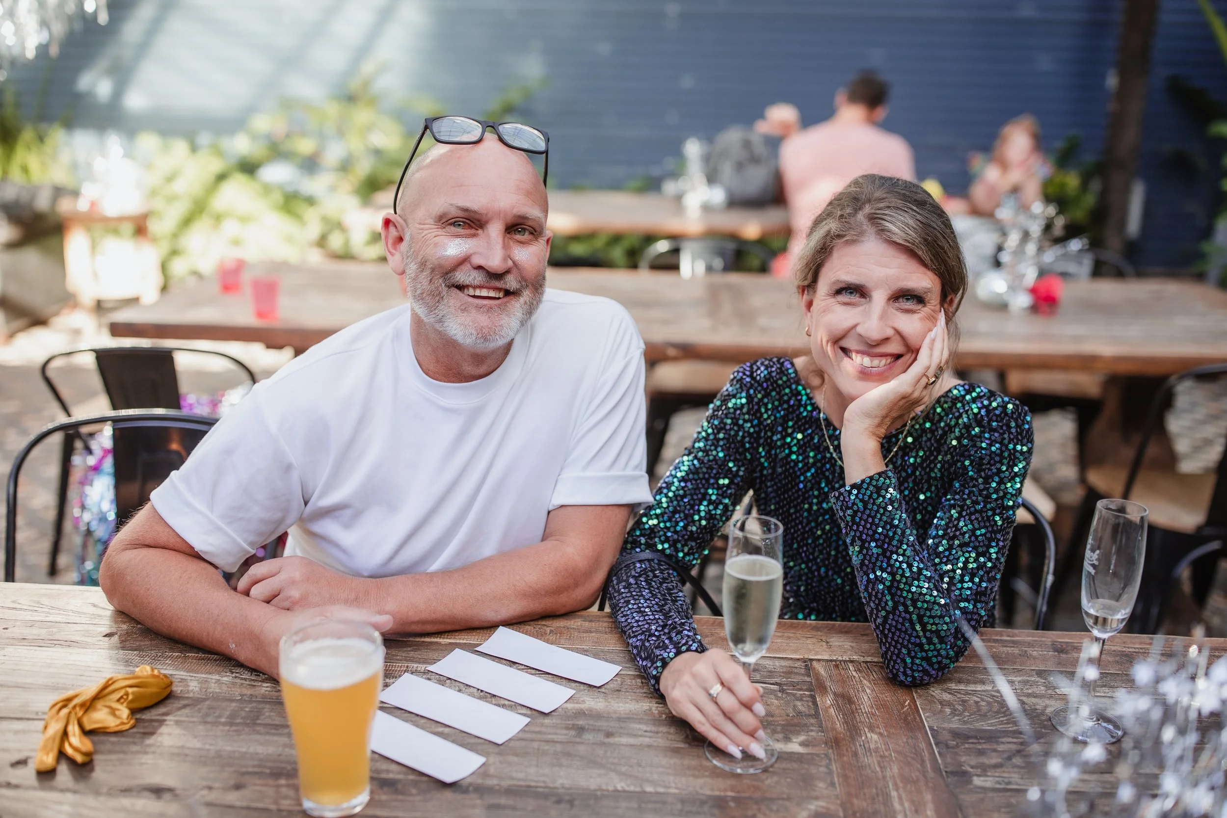 A smiling man and woman sitting at a wooden table outdoors, with drinks and blank notes in front of them, surrounded by other people at a social gathering.