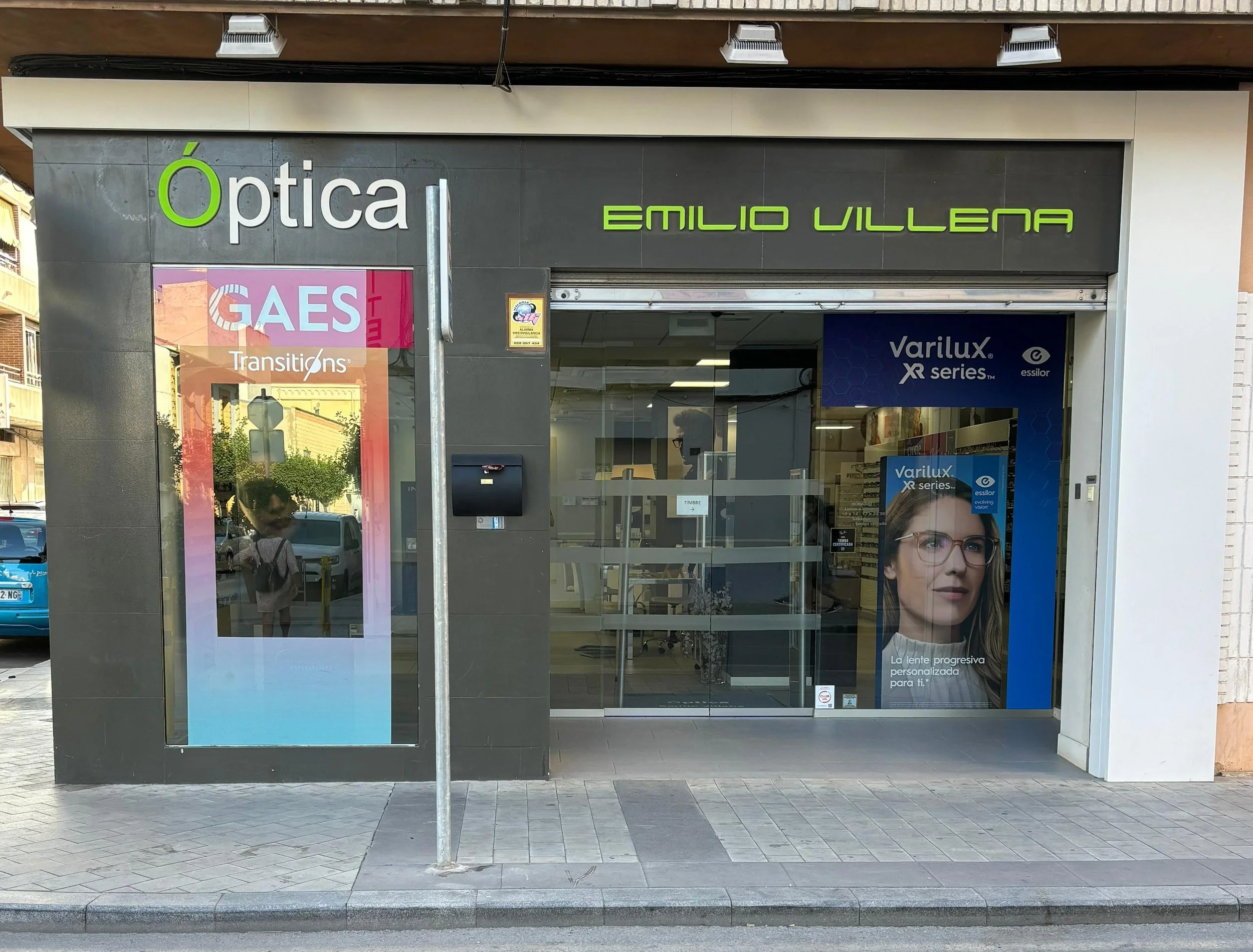 Fachada de Optica Emilio Villena en Almoradi. En la ventana aparece publicidad de GAES ( Audiologia) y de Transitions (Optica). Además de y un anuncio de gafas Varilux XR series con una mujer con gafas.
