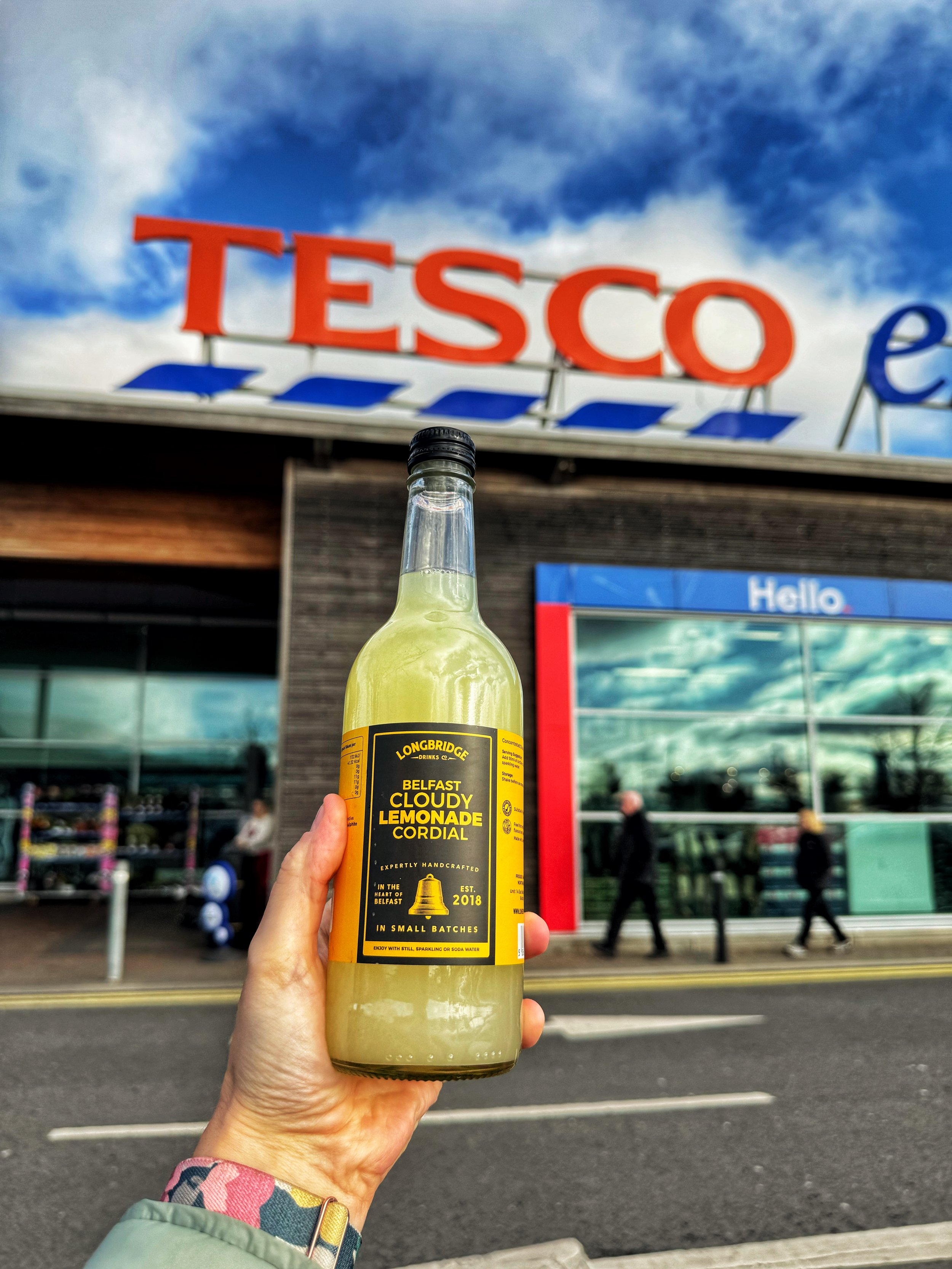 Person holding two bottles of Belfast Cordial drinks, cloudy lemonade and ginger cordial, in front of Tesco Extra parking lot with cars and store sign in the background.