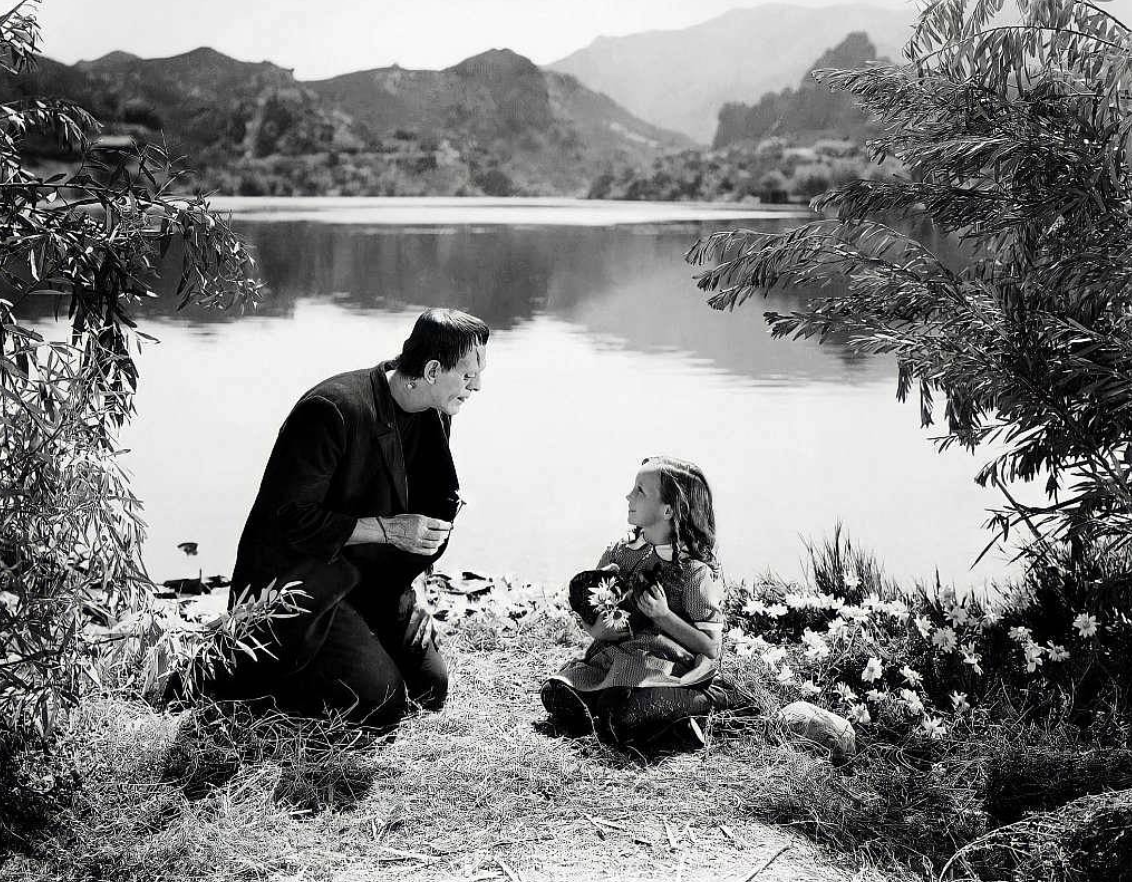a monster crouching next to a little girl at the edge of a lake, she is smiling up at him and there is foliage around them both