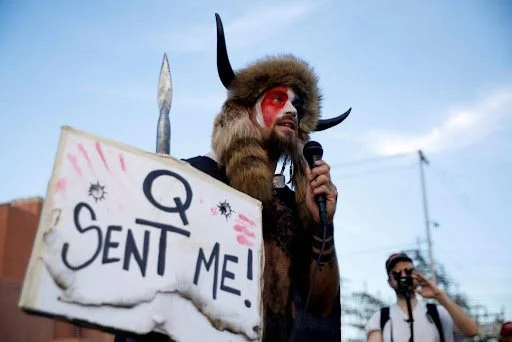 A man wearing red face paint, and a brown hat of animal fur and horns. He is holding a sign stating 'Q SENT ME' and speaking into a microphone