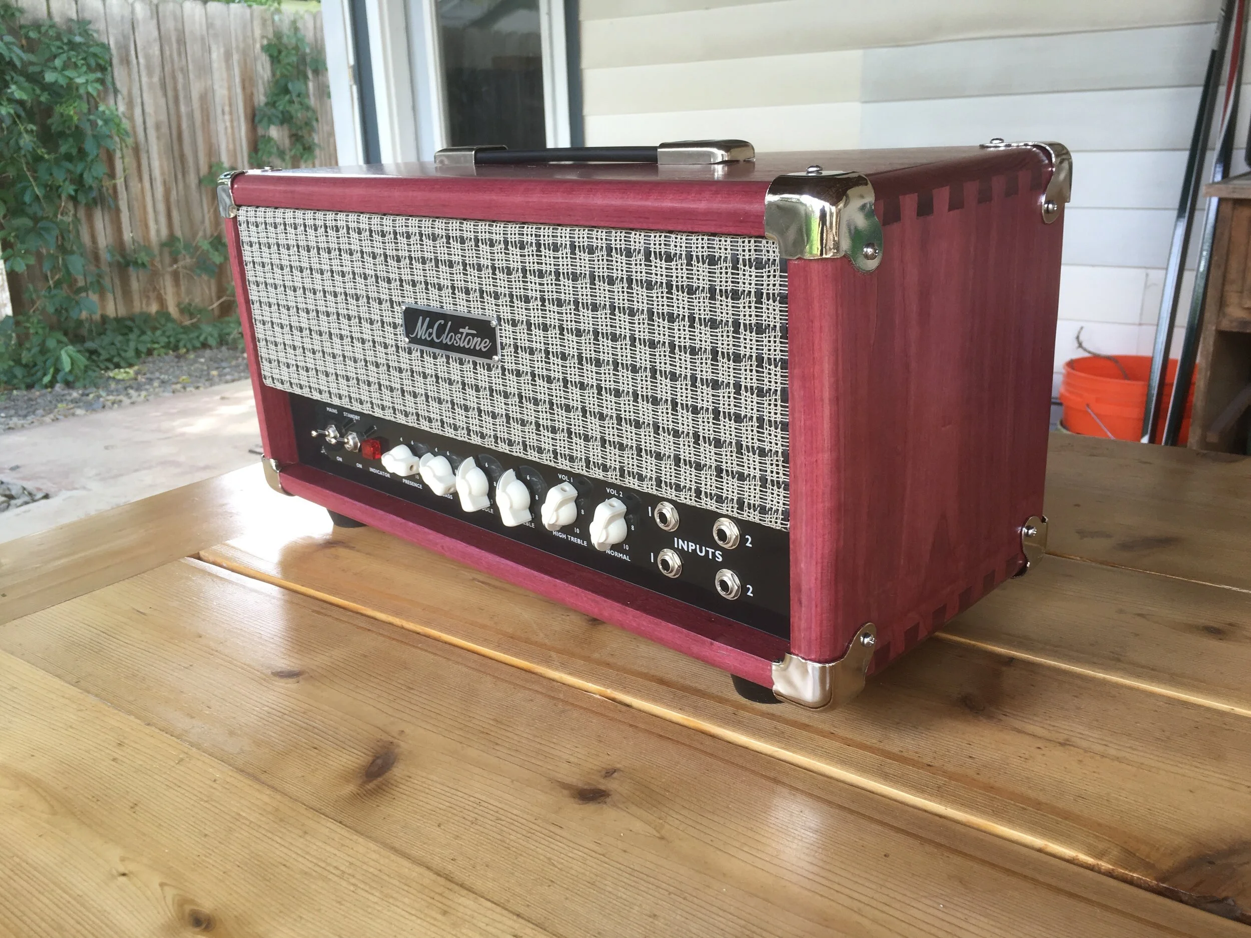 A vintage red McClostone guitar amplifier on a wooden table in a porch or outdoor setting with plants and a white wooden wall in the background.