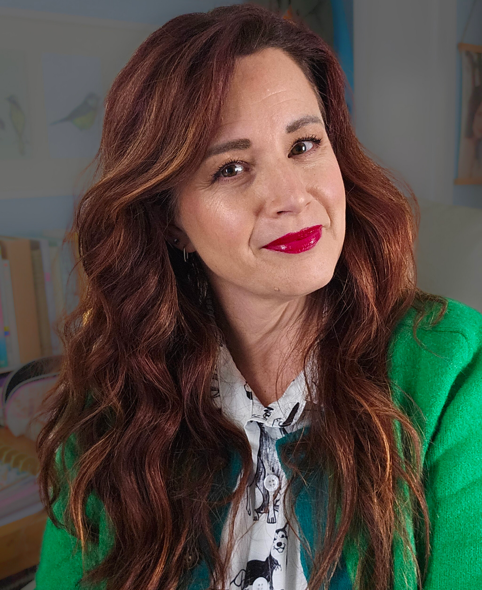 A woman with long, wavy auburn hair and red lipstick smiling, wearing a patterned blouse and a green cardigan, indoors with bookshelf and wall art in the background.