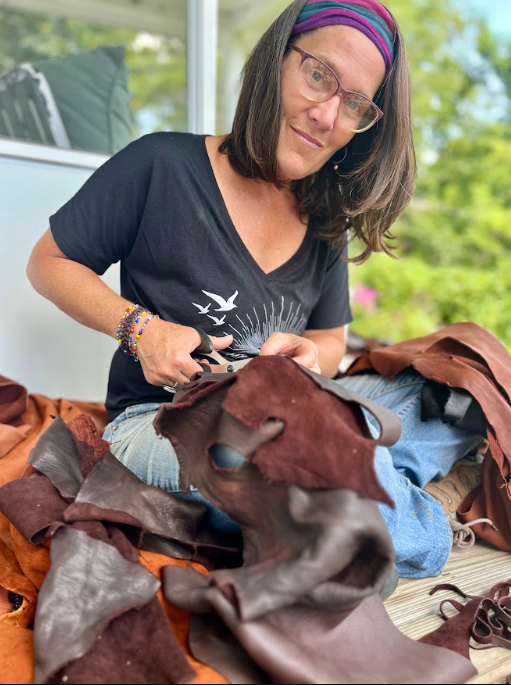 Woman with glasses and a colorful headband sitting outdoors, cutting leather supplies.