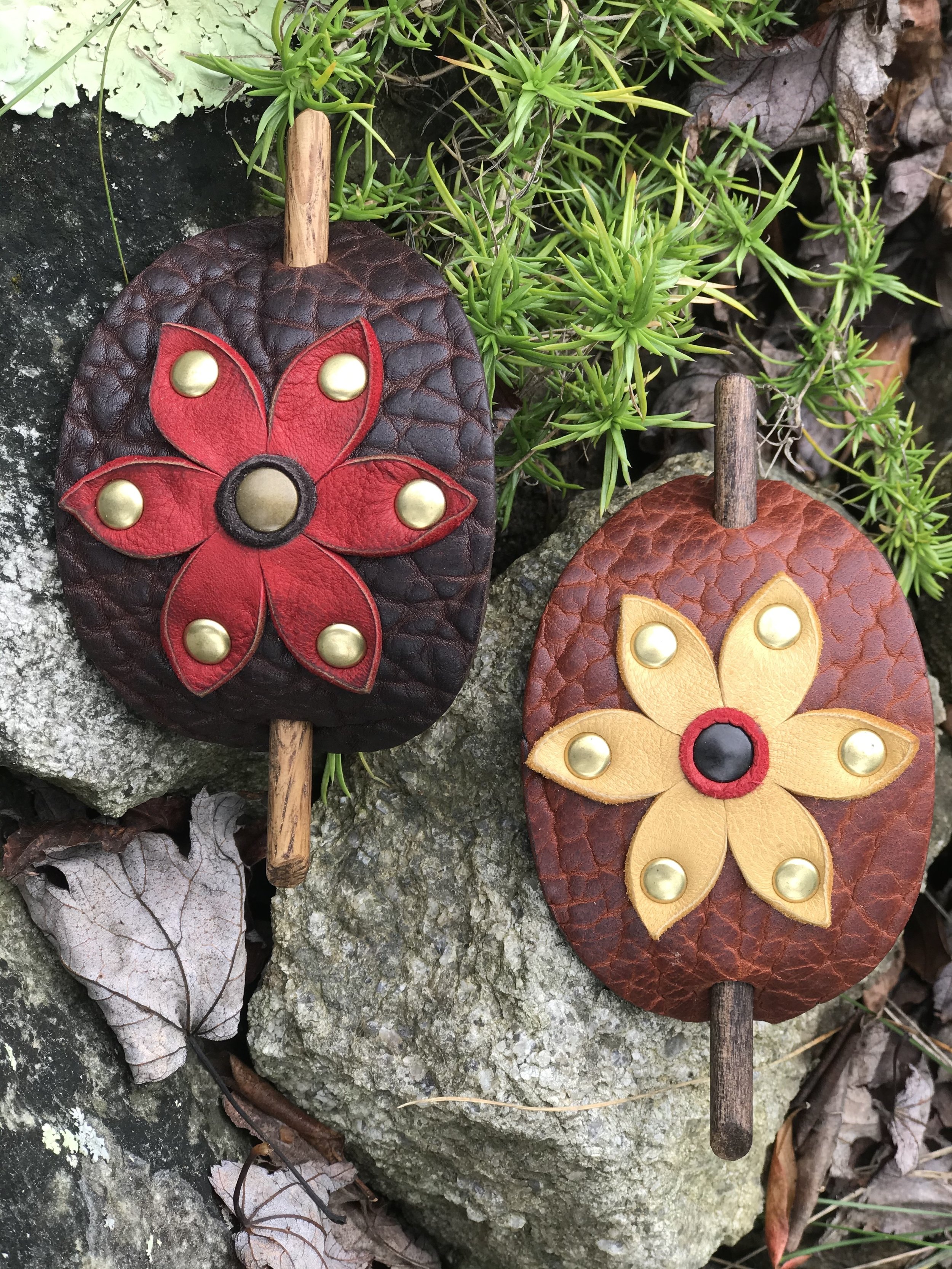 Two handcrafted leather butterfly-shaped decorations with a floral design, set against a background of rocks, leaves, and small green plants.