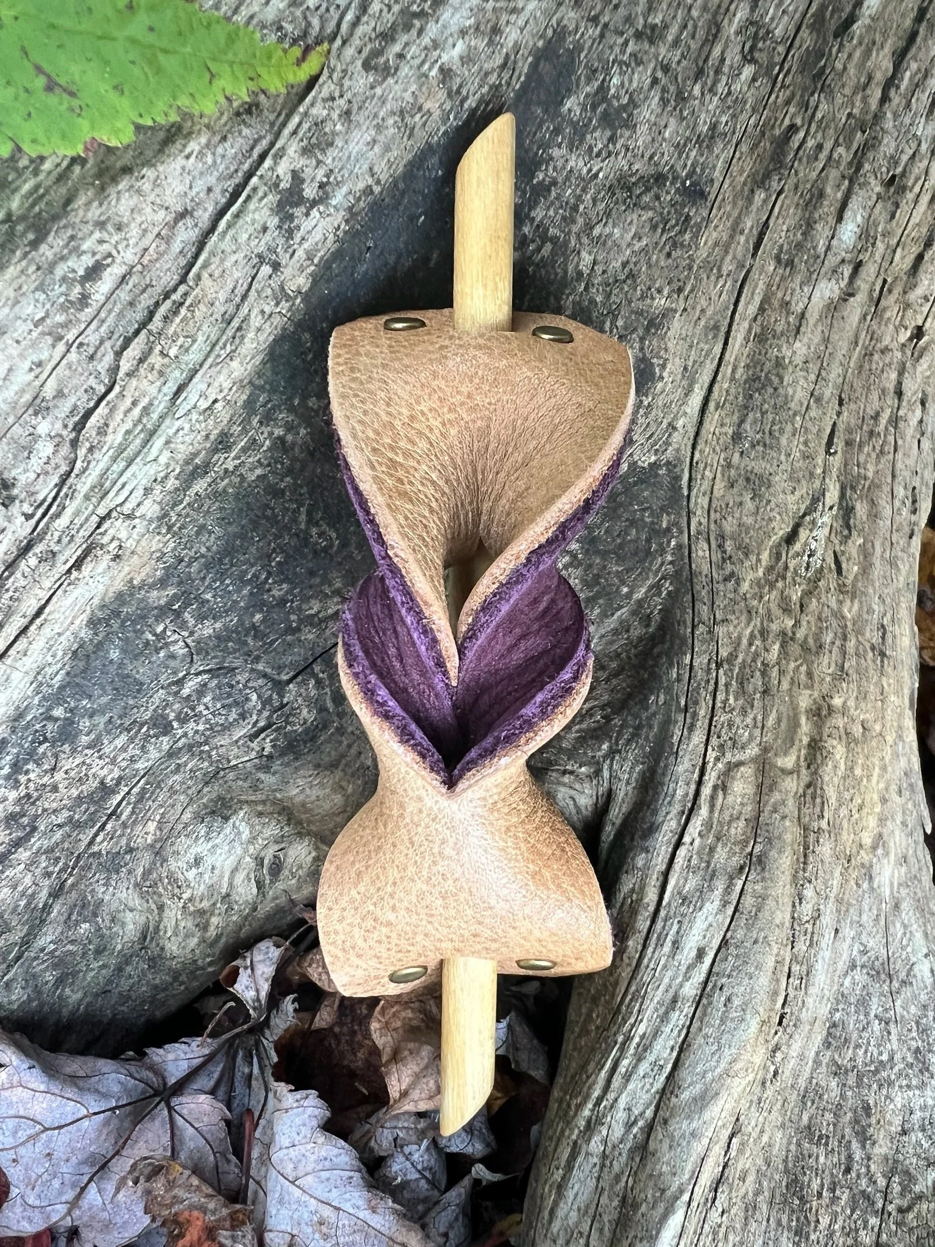 A wooden sculpture resembling a flower with a purple and brown petal design and a beige leaf, mounted on a wooden stick, is placed on a weathered wooden surface with fallen leaves around it.