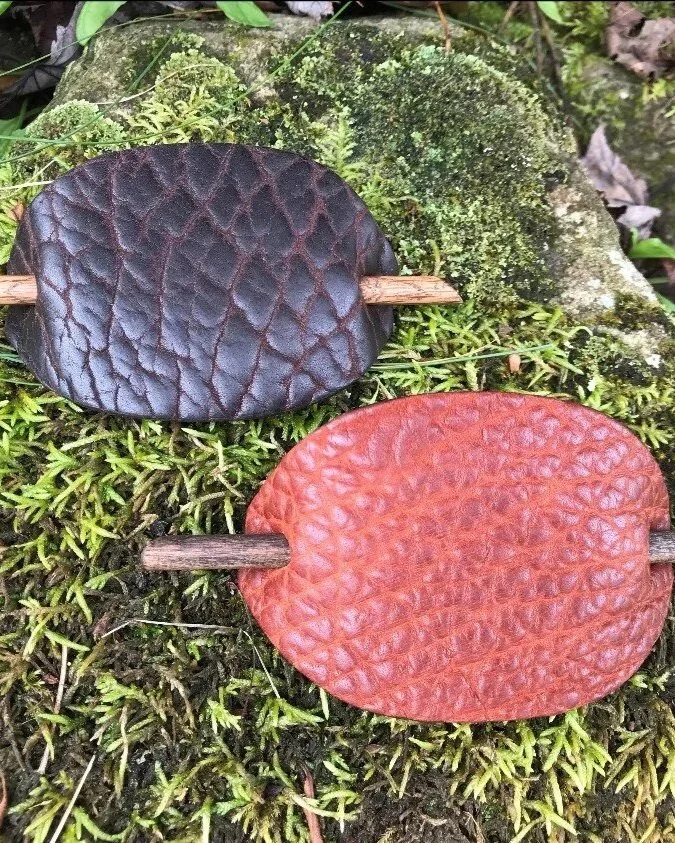 Two leather pads, one dark brown and one reddish-brown, are attached to small wooden sticks and laying on moss-covered ground in a natural setting.