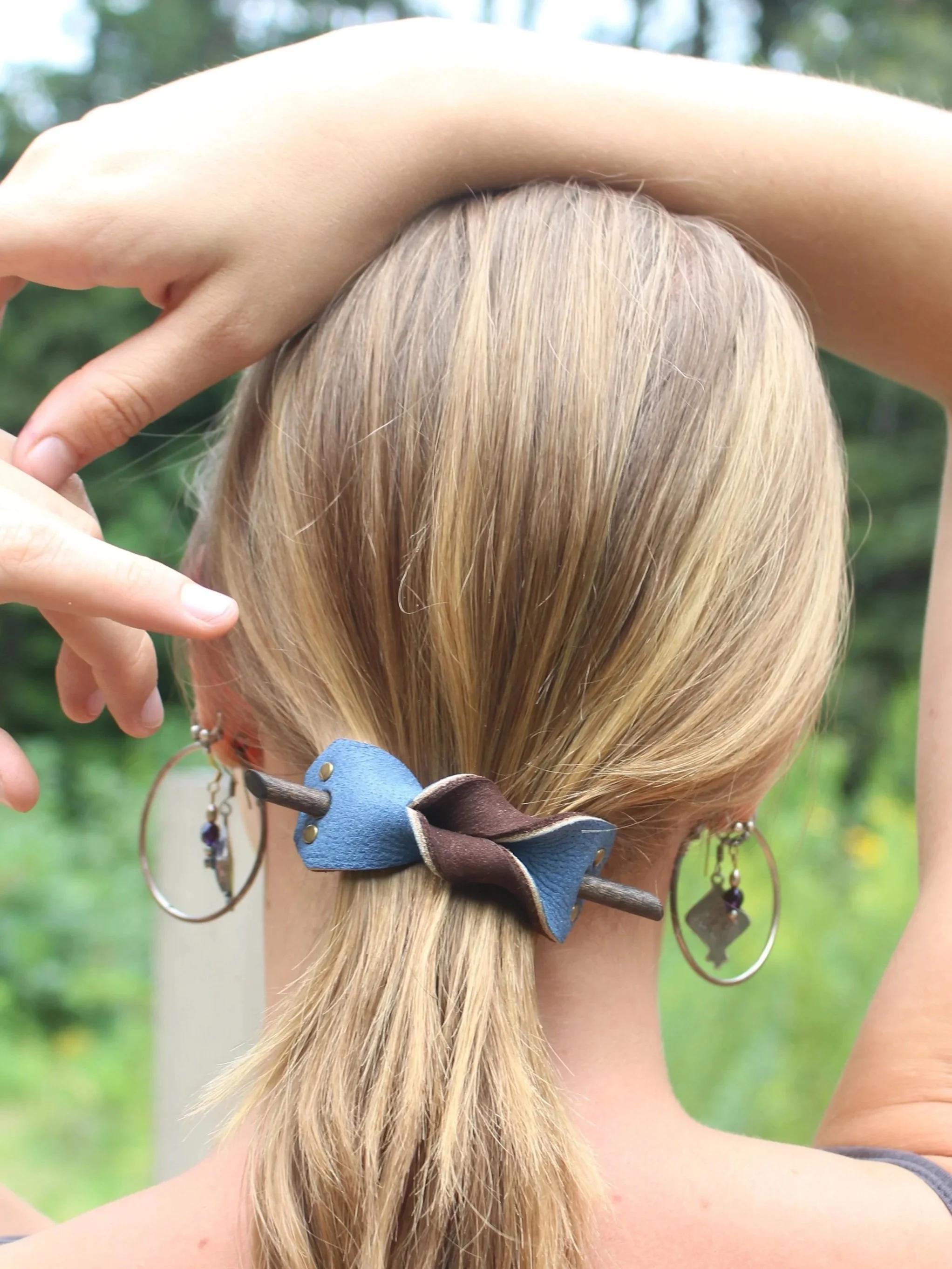 Back of a woman with blonde hair styled in a ponytail secured with a blue leather bow hair tie. She is wearing large hoop earrings and a bracelet with purple beads. One arm is raised across her head, and her background is blurred greenery.